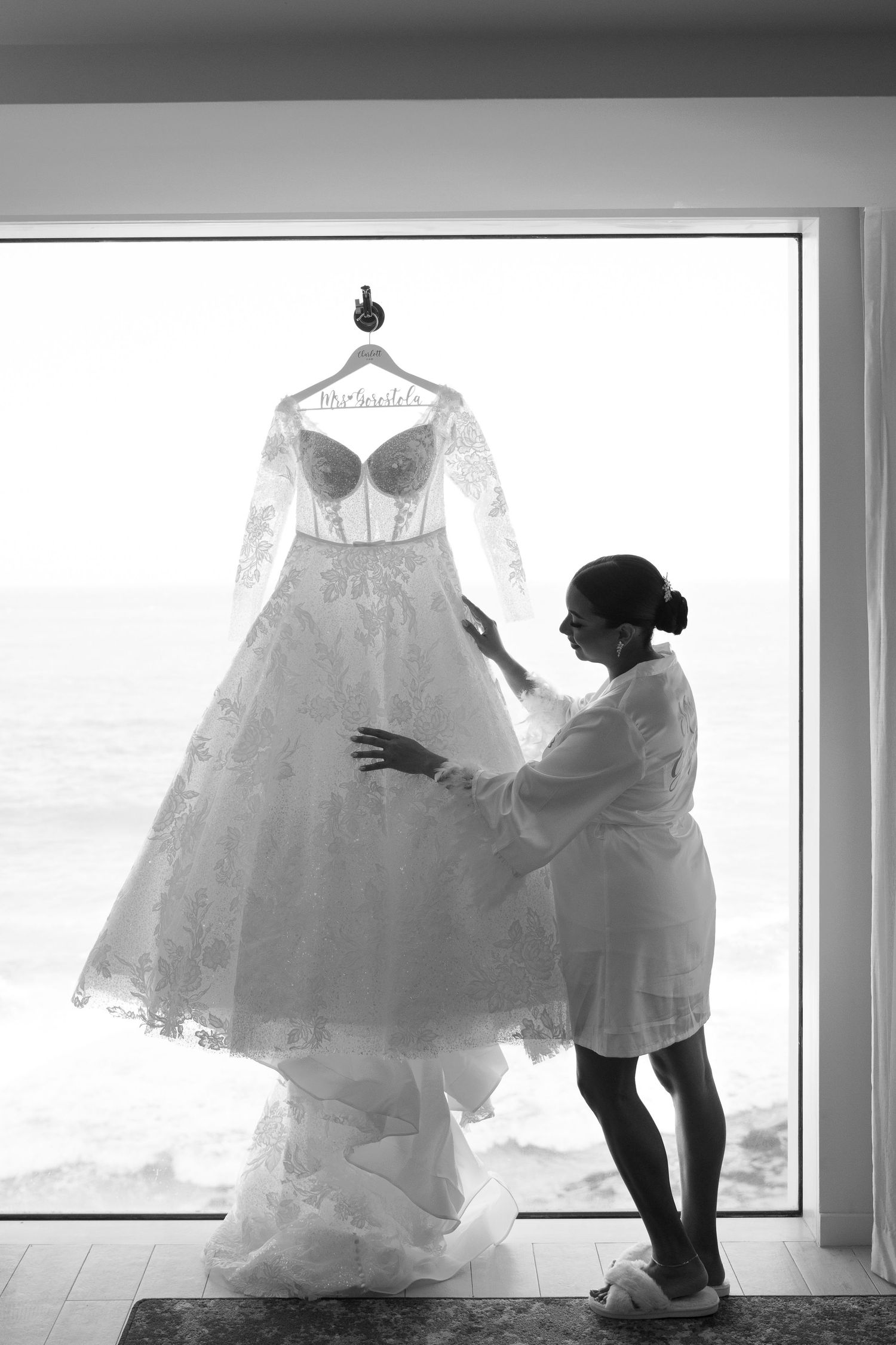 Bride admiring her lace wedding gown hanging by a large ocean-view window in a black and white photo.
