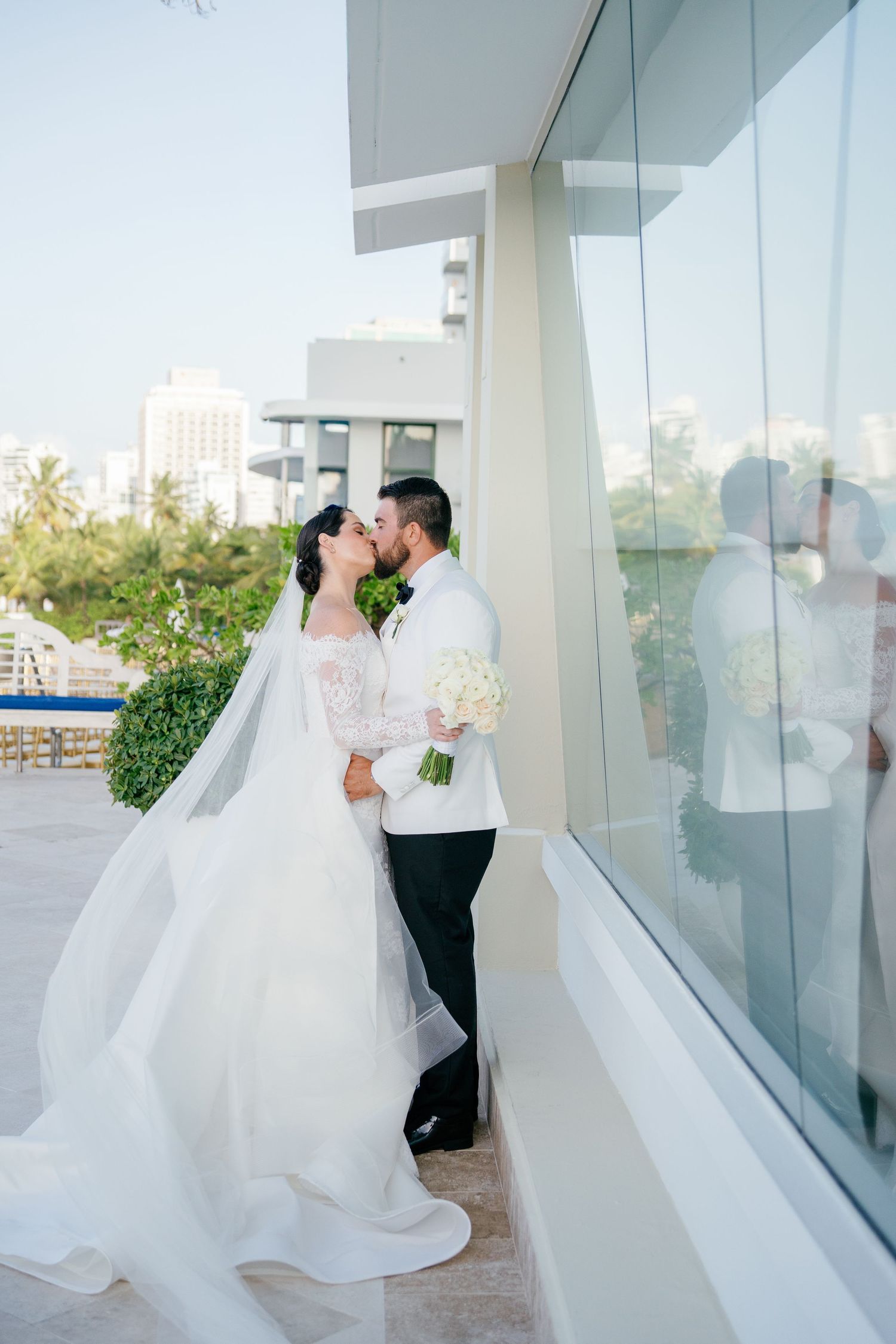 Bride and groom share a kiss by a glass wall, her veil flowing, holding a white bouquet at an outdoor venue.