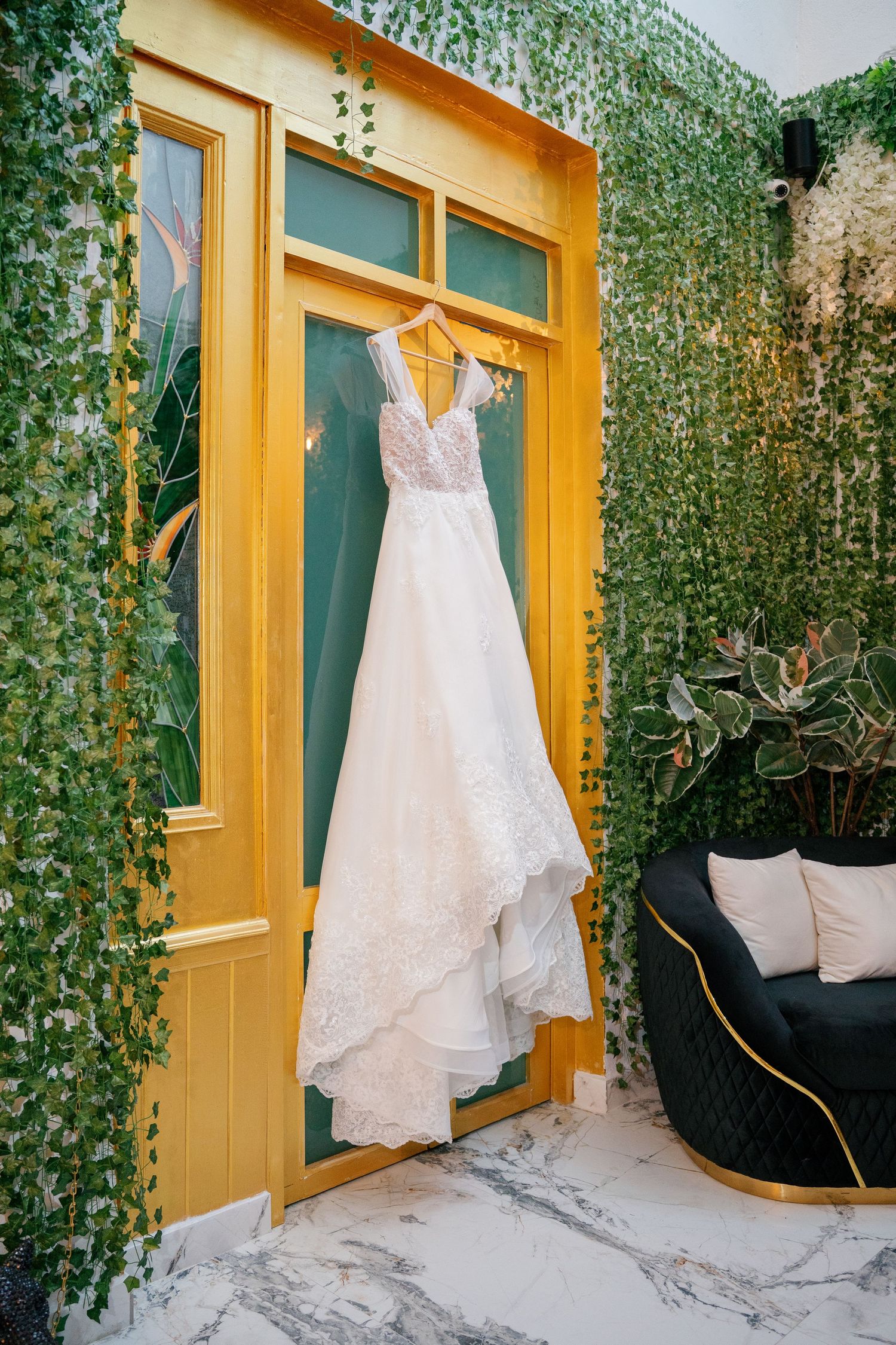 White lace wedding dress hanging on yellow door frame surrounded by green ivy wall with black velvet chair nearby.