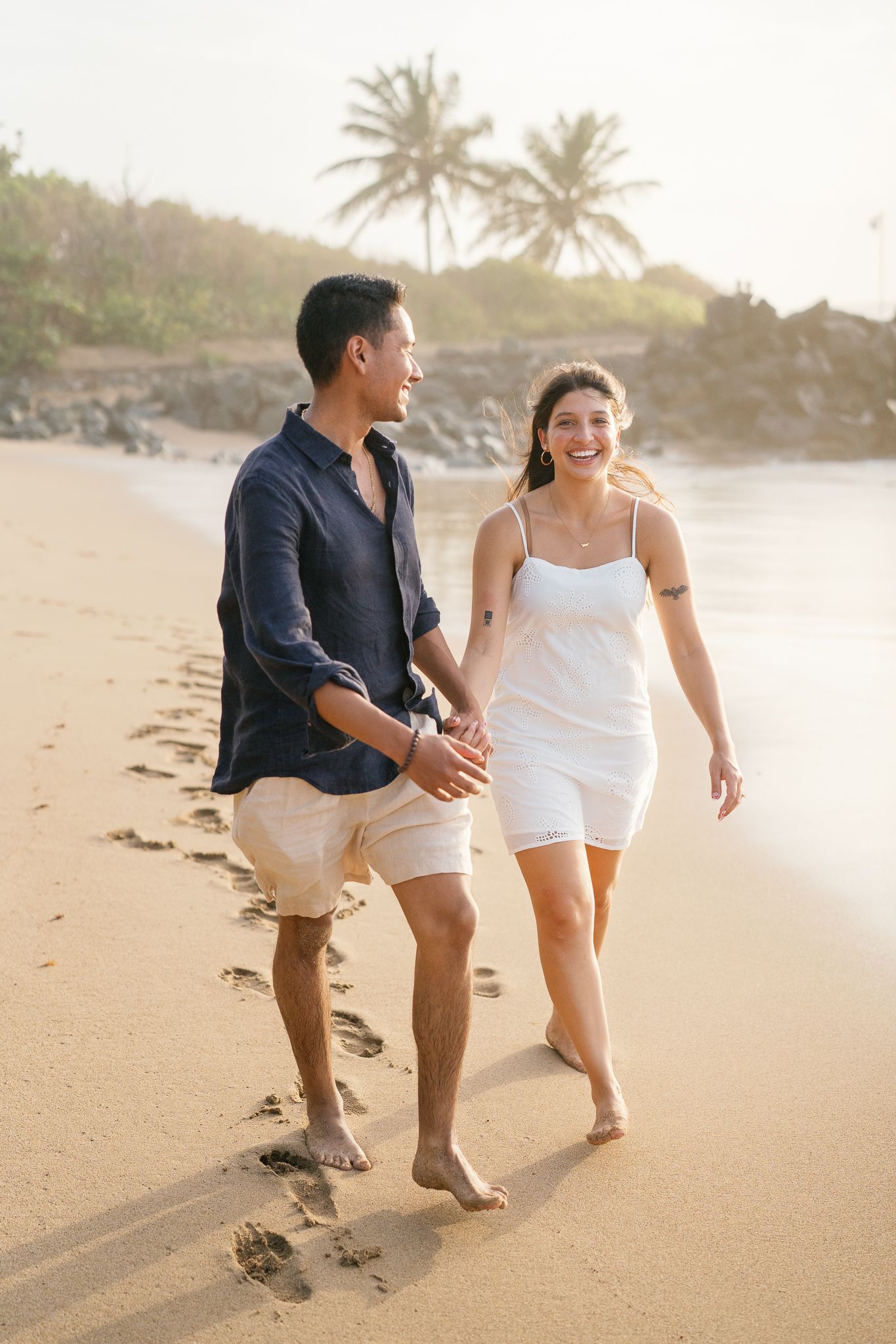 A couple shares a playful moment walking along a tropical beach at sunset with palm trees in the background.