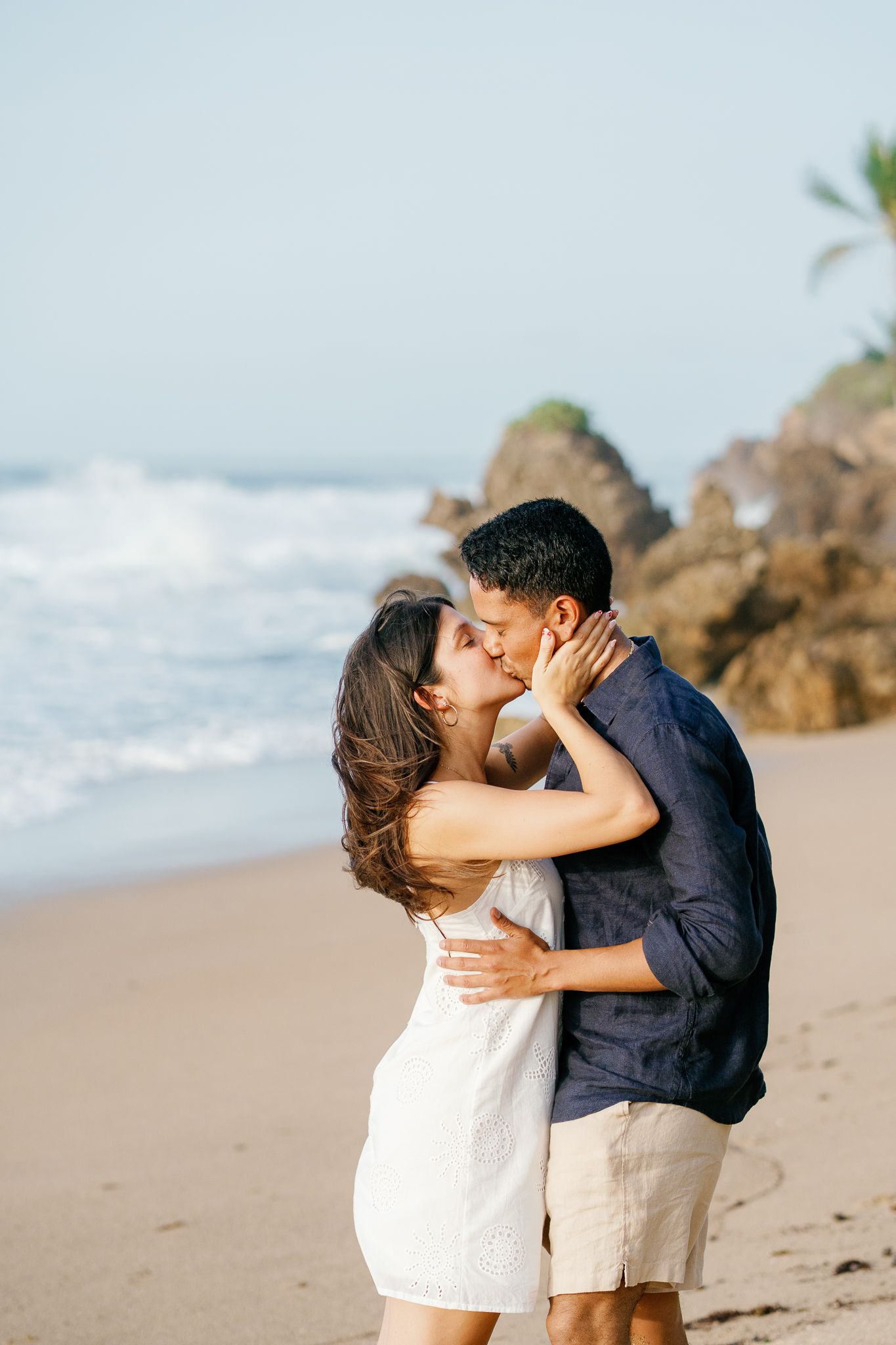 A romantic couple shares a tender kiss on a sandy beach with ocean waves and rocks in the background.