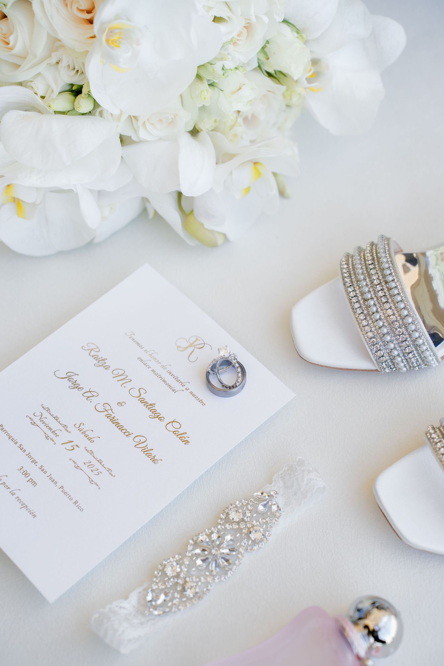 Elegant wedding flat lay with white floral bouquet, crystal bridal shoes, diamond rings, rhinestone belt, and gold script invitation.
