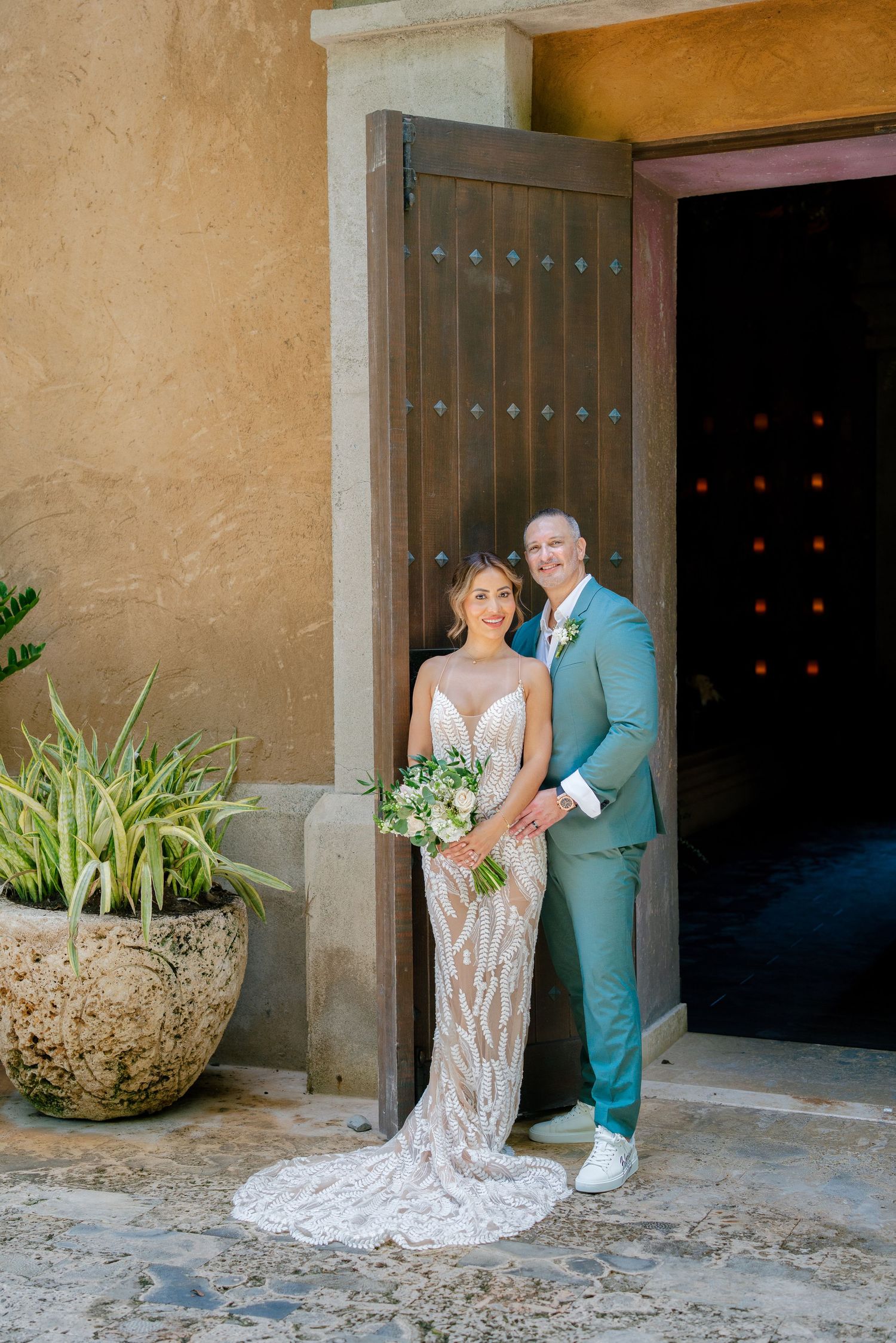 Wedding couple posing by rustic wooden doors and stone walls in tropical setting.