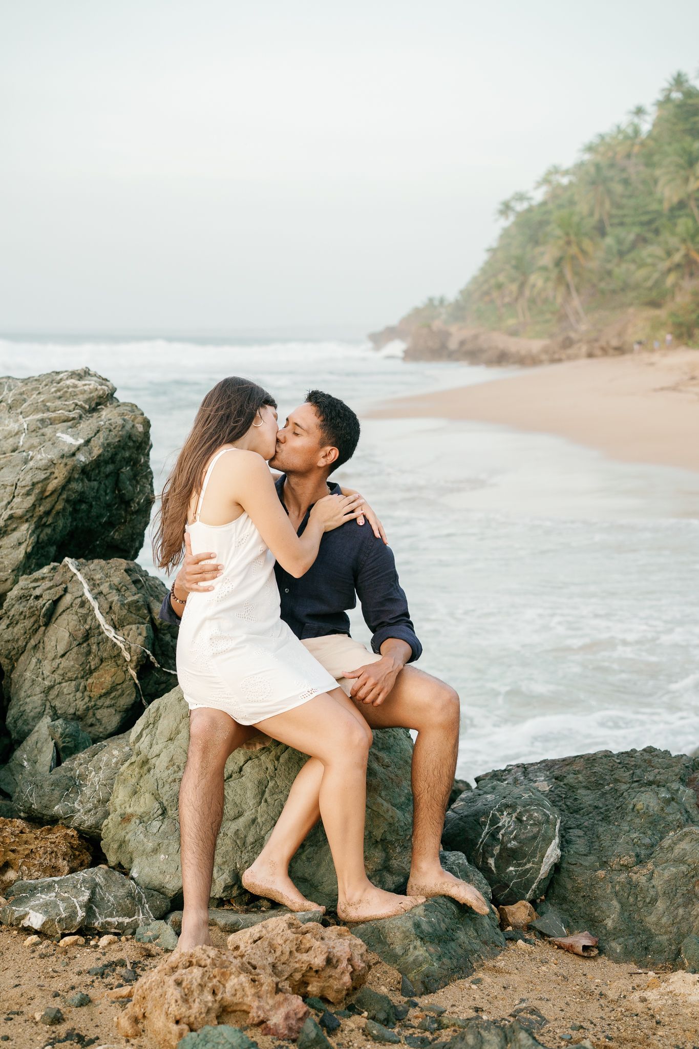 Romantic couple sharing an intimate moment on rocky beach with crashing waves and palm trees in background.