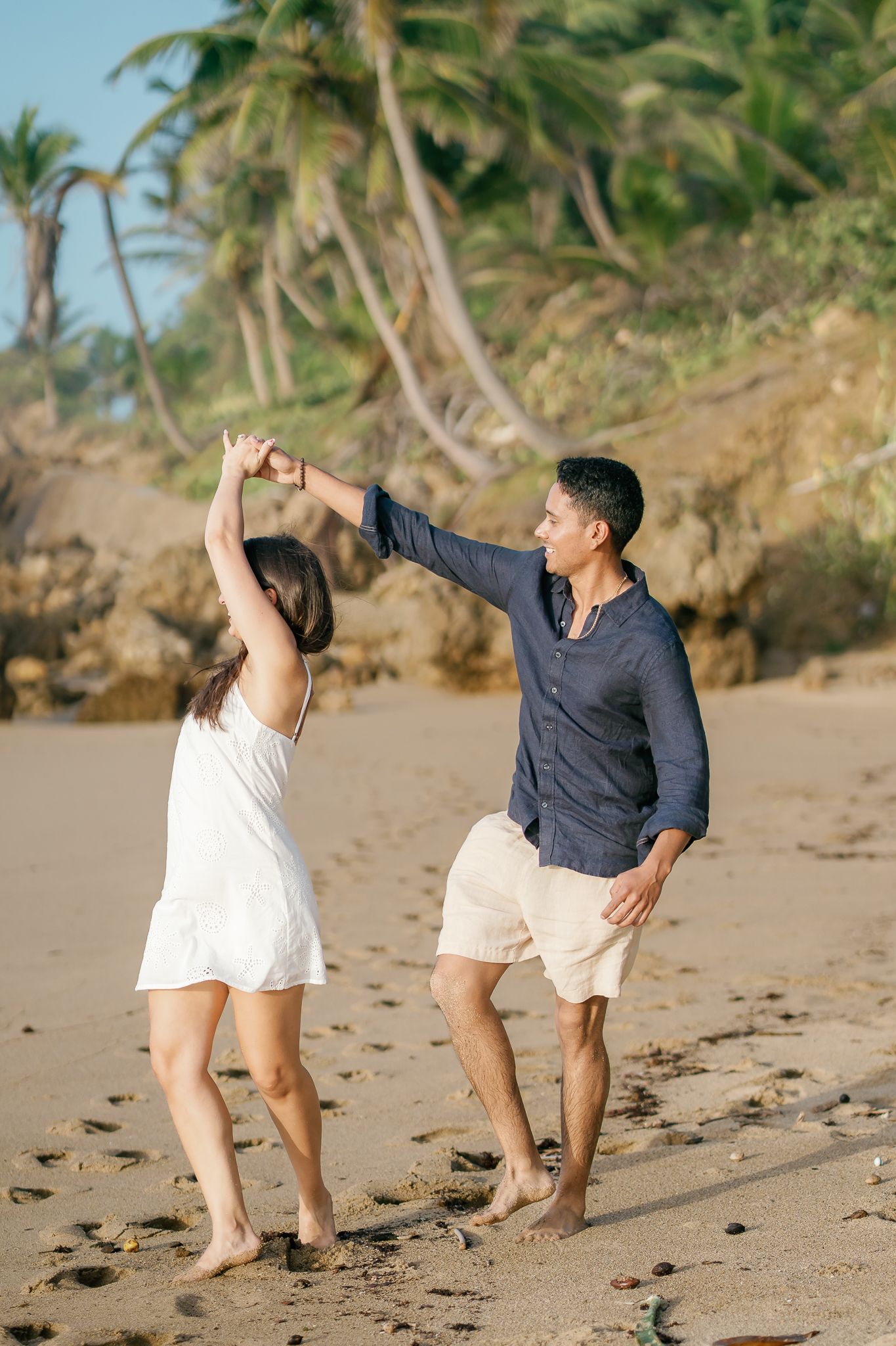 A couple dances playfully together on a sandy tropical beach with palm trees in the background during golden hour.
