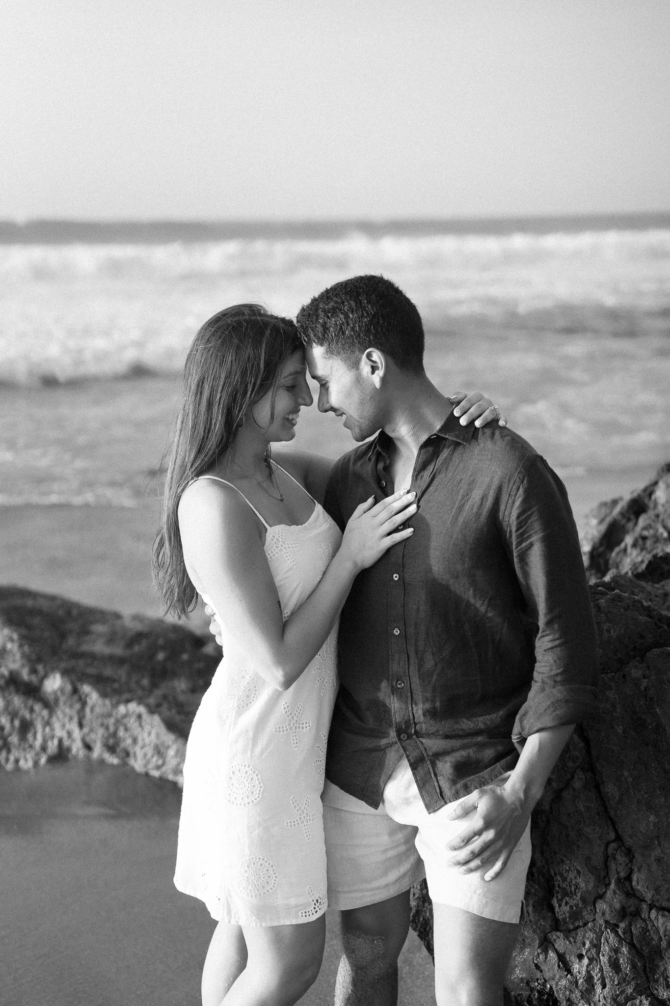 A romantic couple shares an intimate moment on a rocky beach with ocean waves in the background.