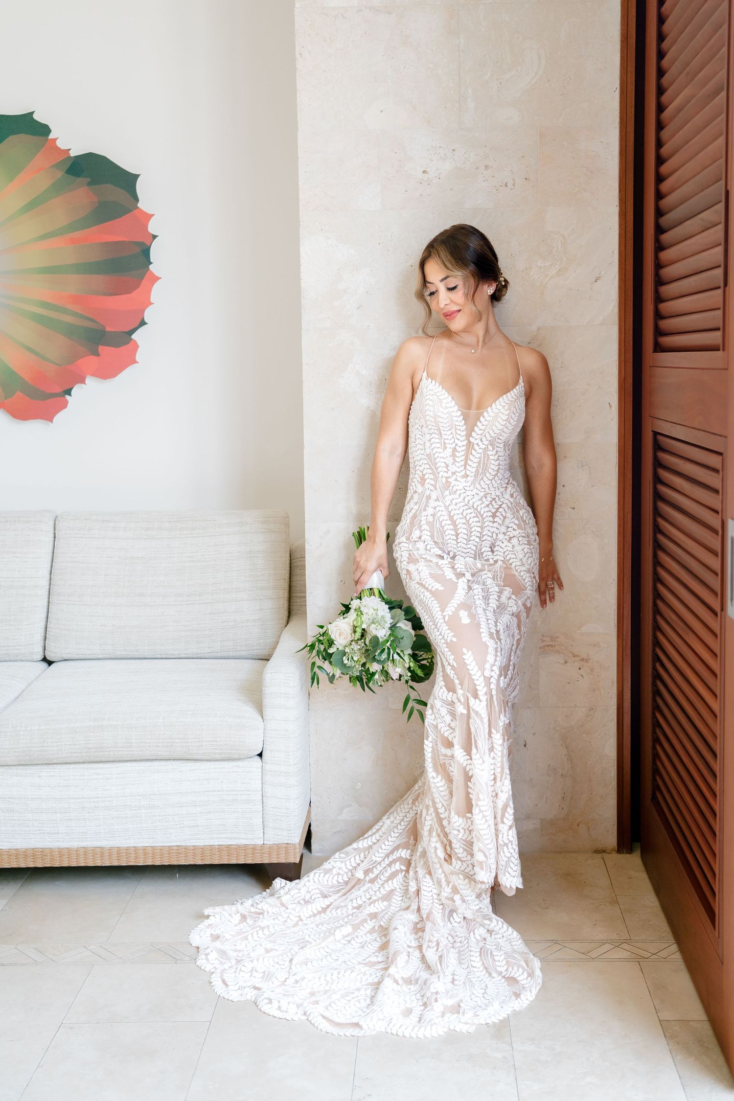 Full-length view of wedding dress with train and green bouquet against neutral wall.