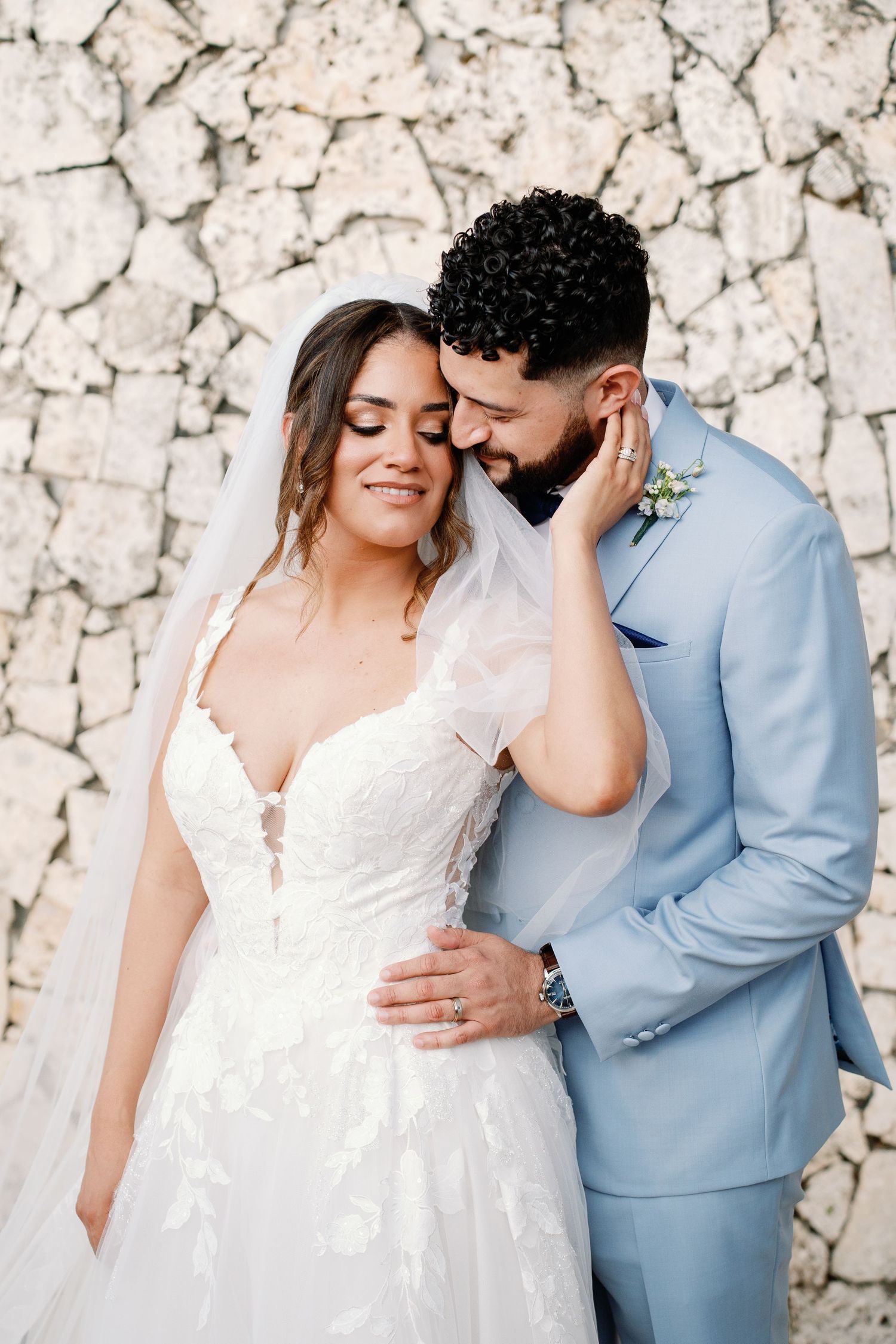 Bride in lace gown and veil embraces bearded groom in light blue suit against a stone wall backdrop.