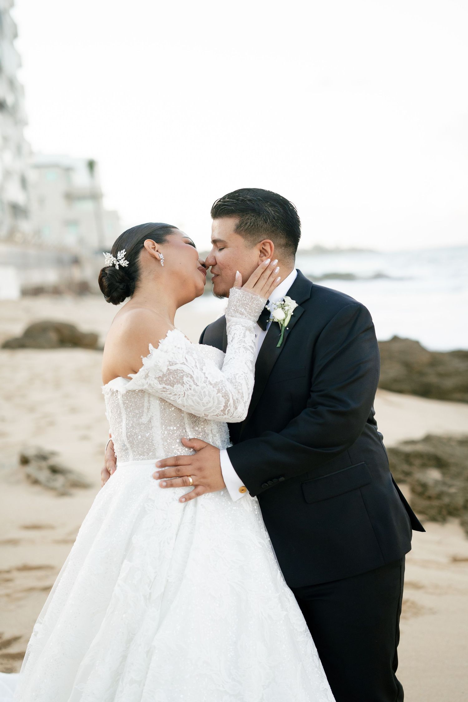 Bride and groom share a romantic beach kiss, she in lace off-shoulder gown, he in black tuxedo.