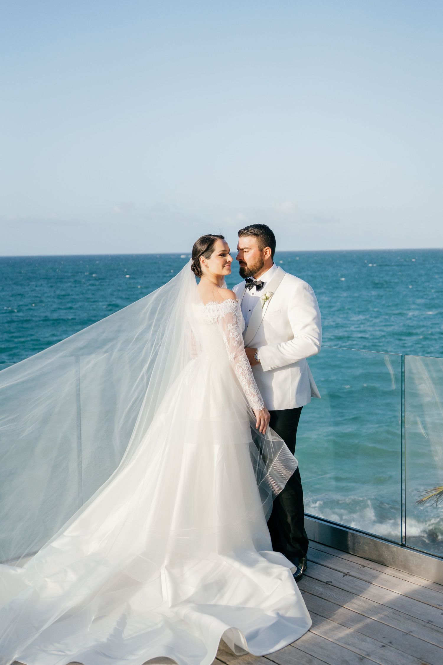 Bride in flowing lace gown with dramatic veil and groom in white tuxedo embrace on oceanfront terrace.