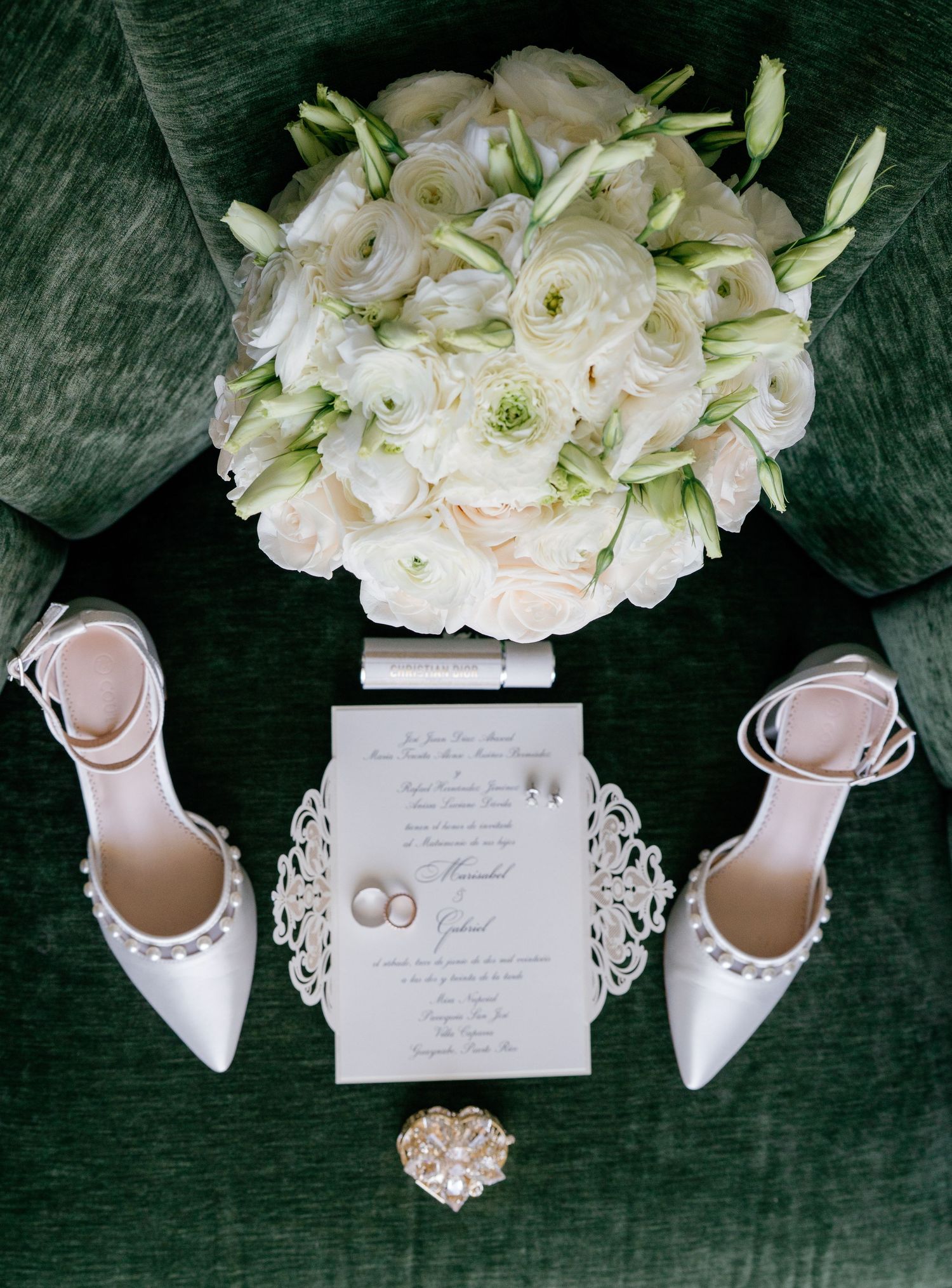 Elegant wedding flat lay with white floral bouquet, pearl heels, rings, and ornate invitation on green velvet chair.