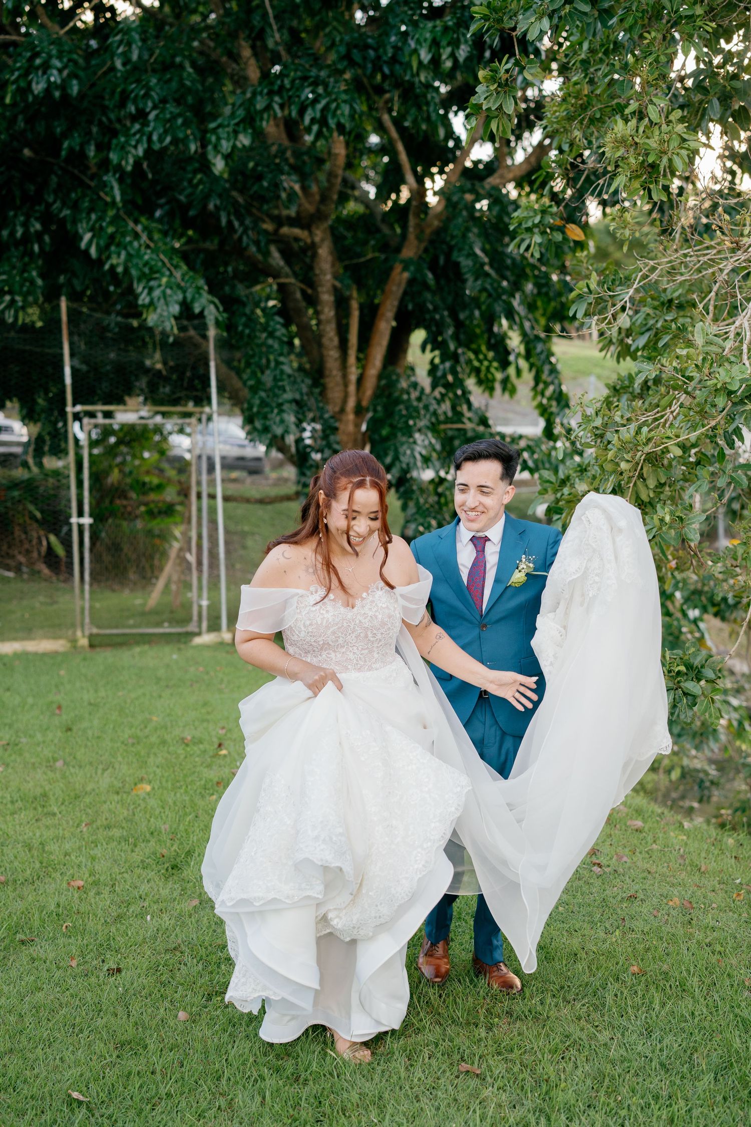Groom in blue suit helps bride in off-shoulder lace gown walk across green lawn during outdoor wedding celebration.