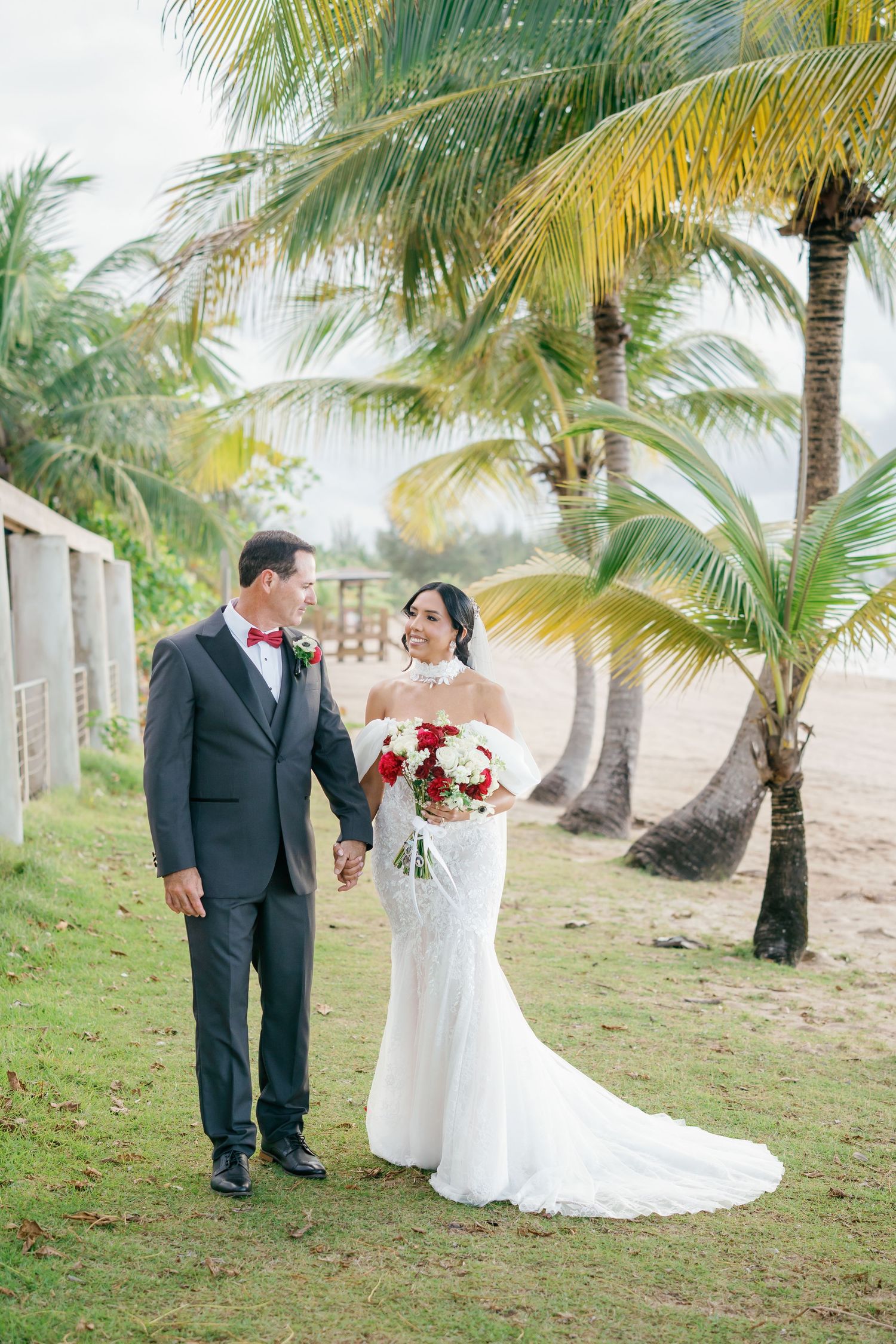 Bride and groom hold hands near palm trees on a tropical beach, she in a white gown with red bouquet, he in a gray suit.