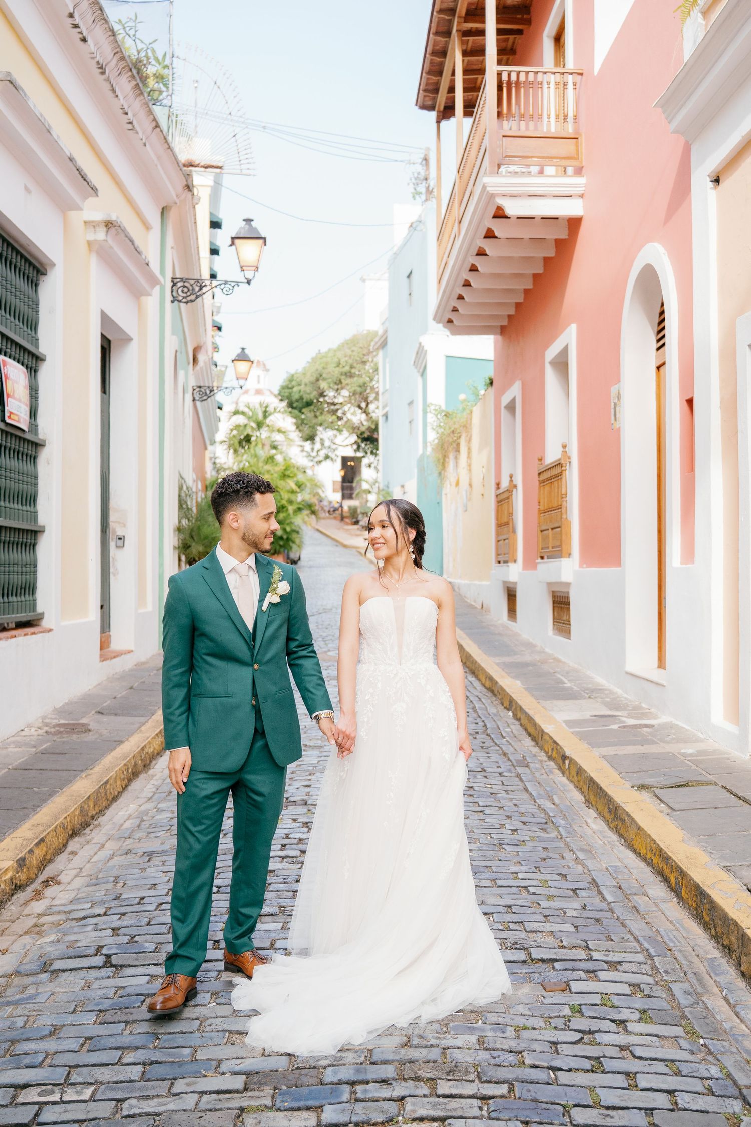 Bride and groom holding hands on colorful cobblestone street in Old San Juan, Puerto Rico, groom in green suit.
