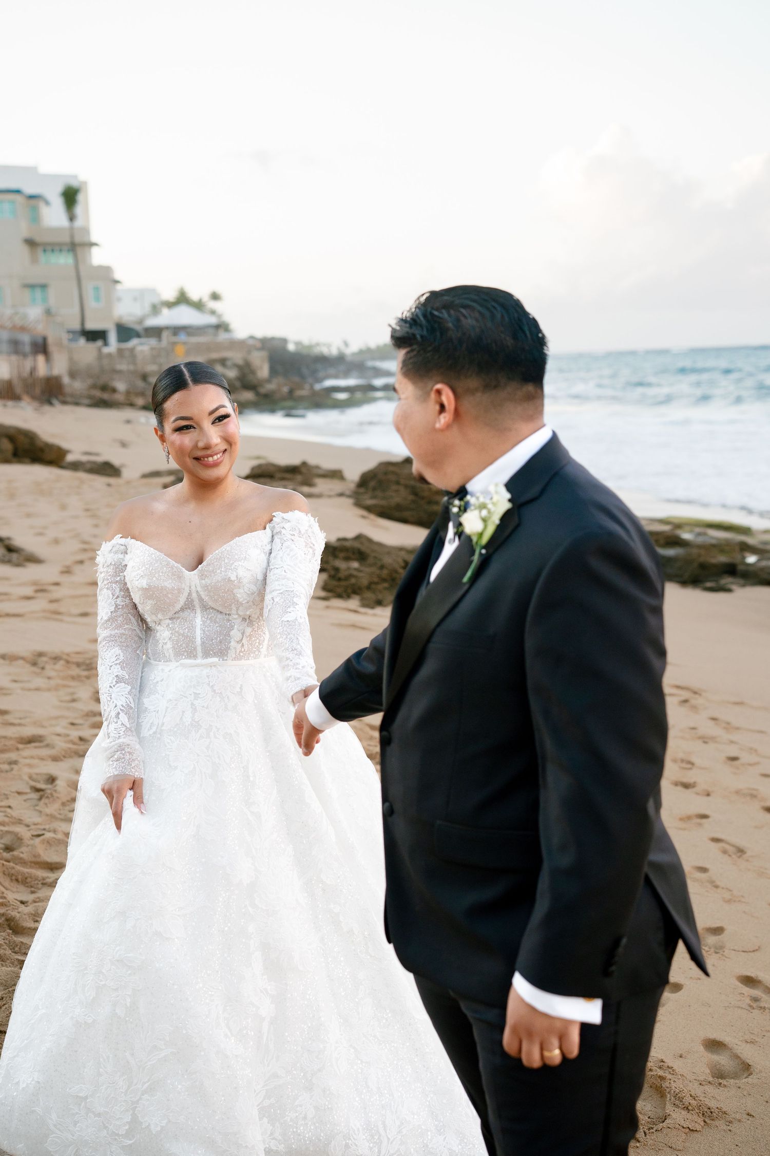 Bride in lace off-shoulder gown holds groom's hand on sandy beach, ocean waves behind them.