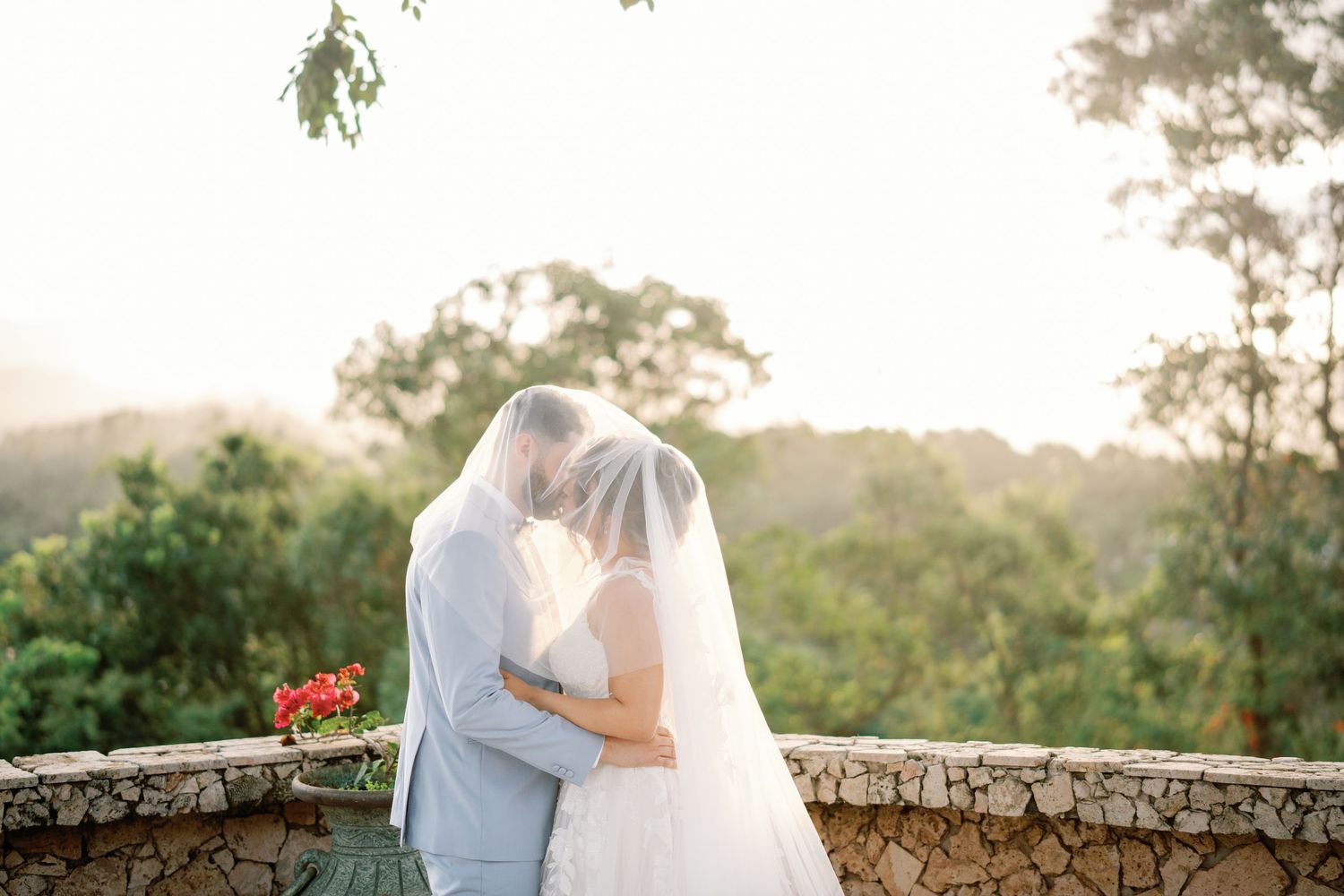 Bride and groom share a kiss under a veil at an outdoor wedding venue with lush green trees in the background.
