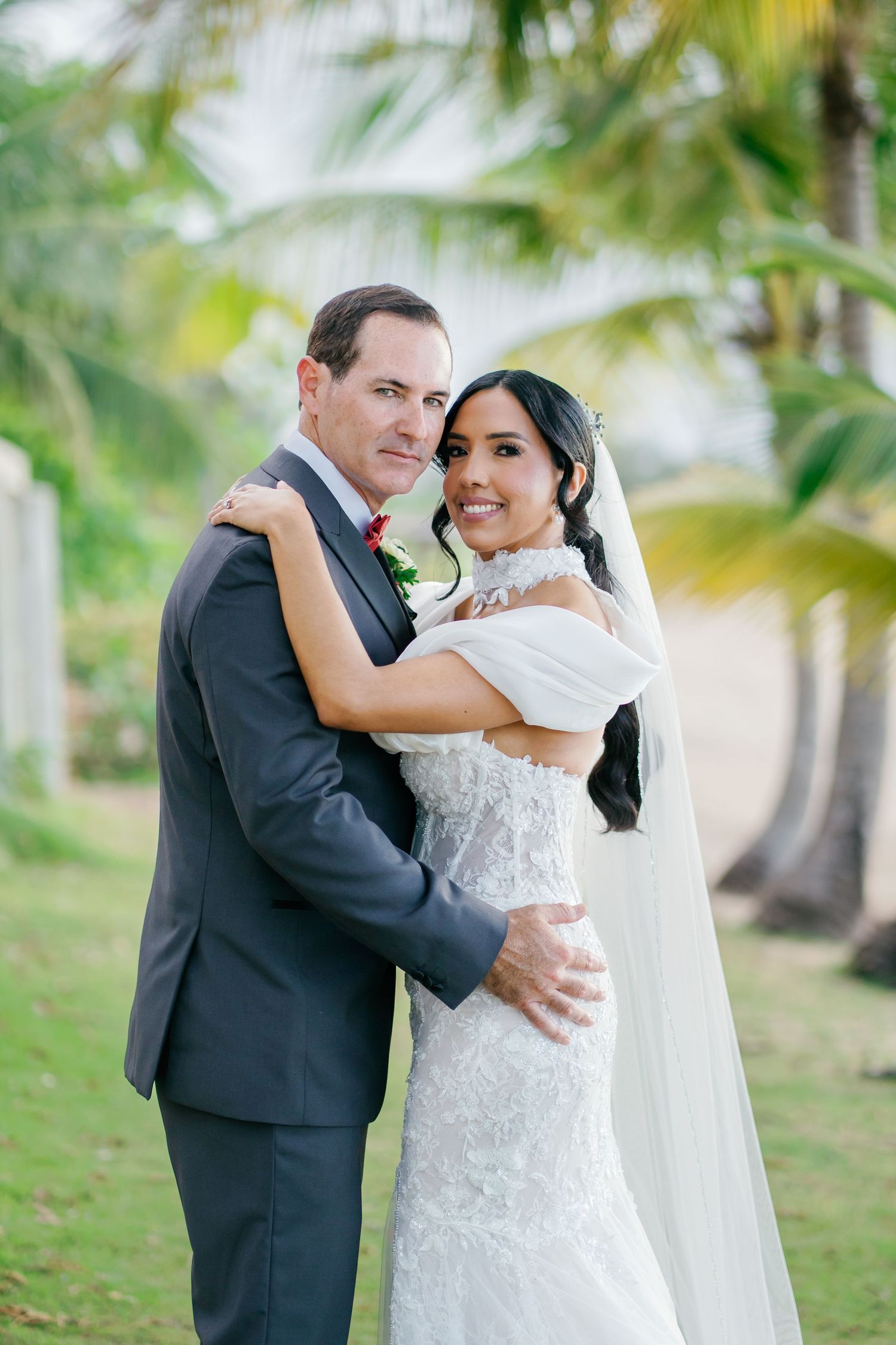 Bride and groom embracing outdoors near palm trees, bride in lace gown with veil, groom in gray suit.