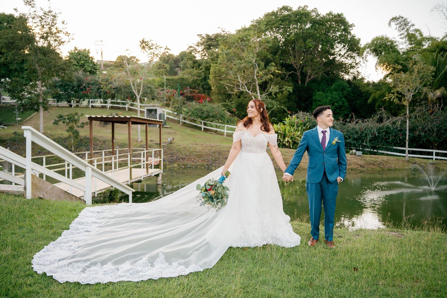 Bride in flowing white gown and groom in blue suit hold hands in lush garden with pond and wooden bridge backdrop.