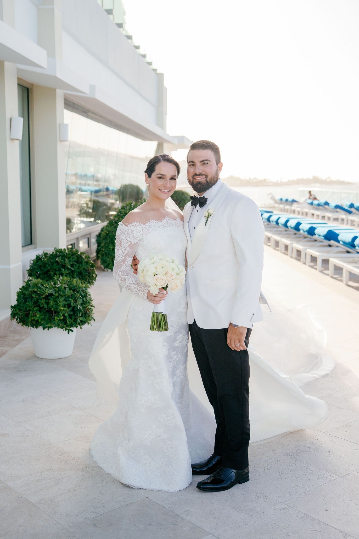 Bride and groom pose together on a waterfront terrace, she in lace gown with bouquet, he in white tuxedo jacket.