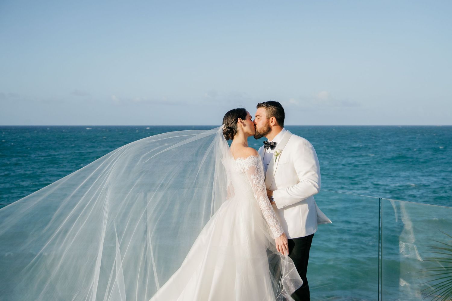 Bride and groom share a kiss on oceanfront terrace, her veil flowing in the breeze, wearing white gown and tuxedo.
