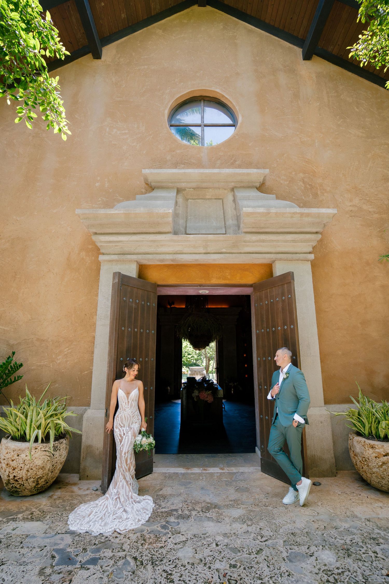 A couple poses at the entrance of a Spanish mission-style church with stucco walls and round window above the doorway.