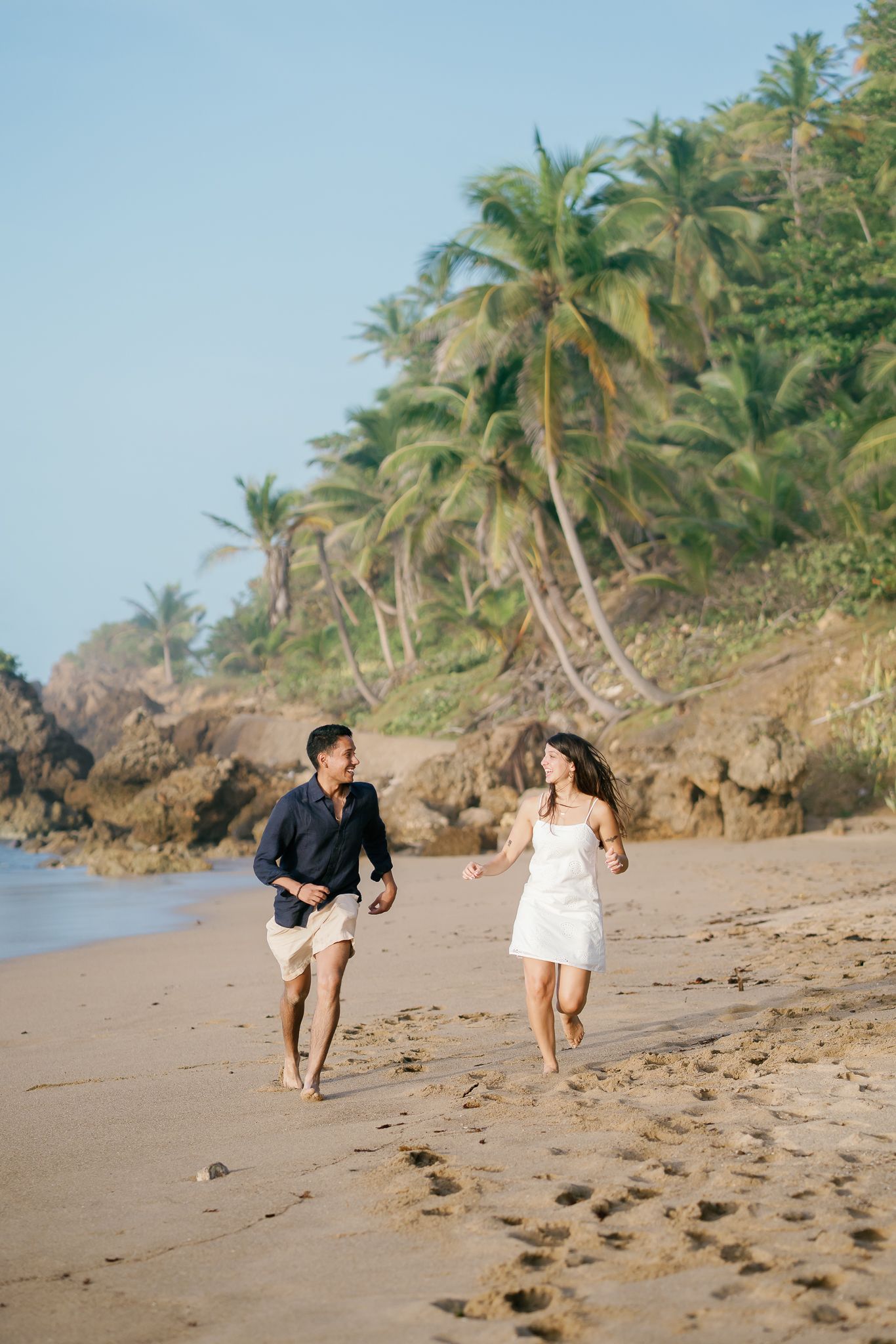 A couple runs joyfully along a tropical beach with palm trees and rocky cliffs in the background.