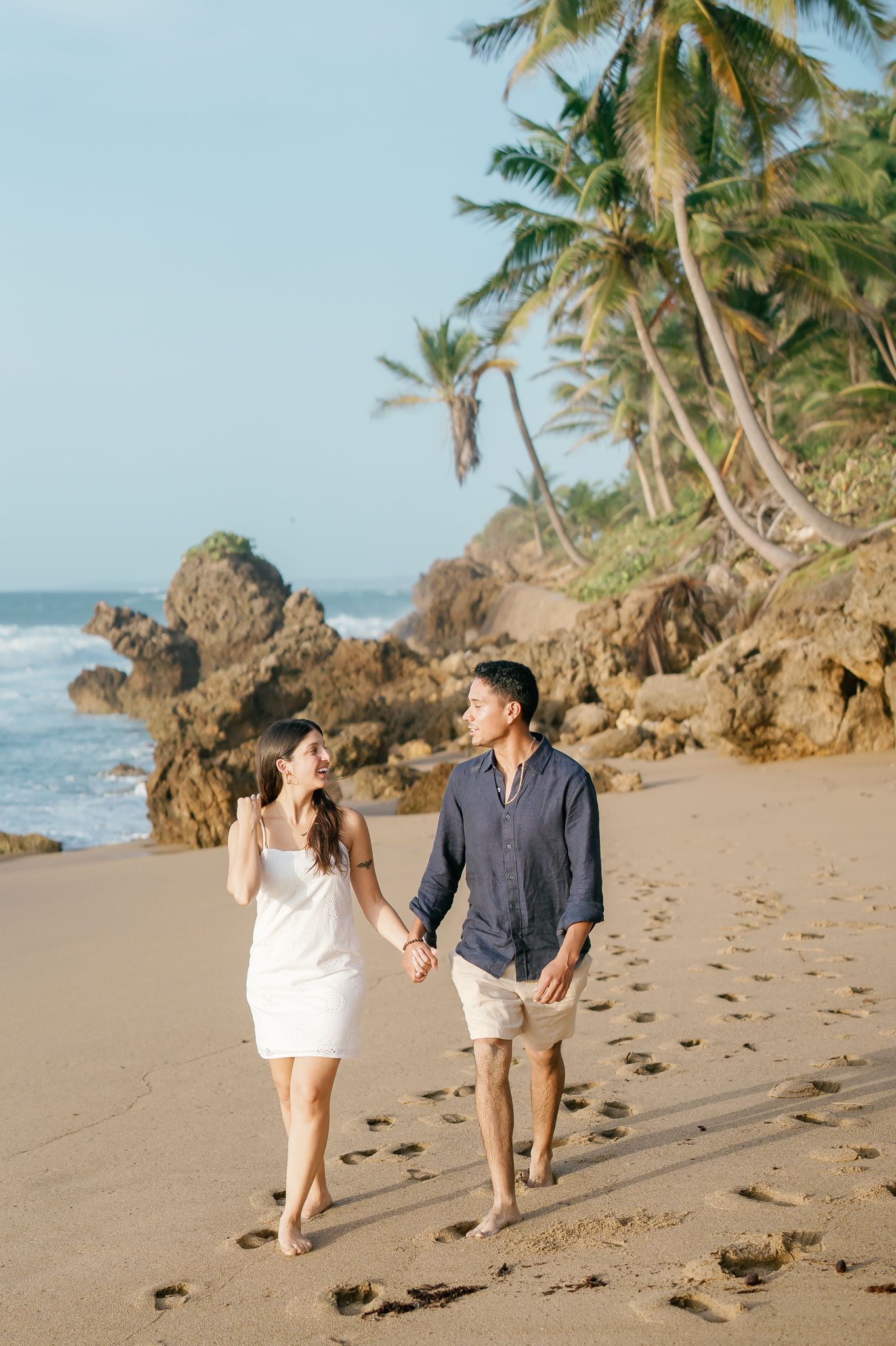 A couple walks along a tropical beach with palm trees and rocky outcrops in Puerto Rico.