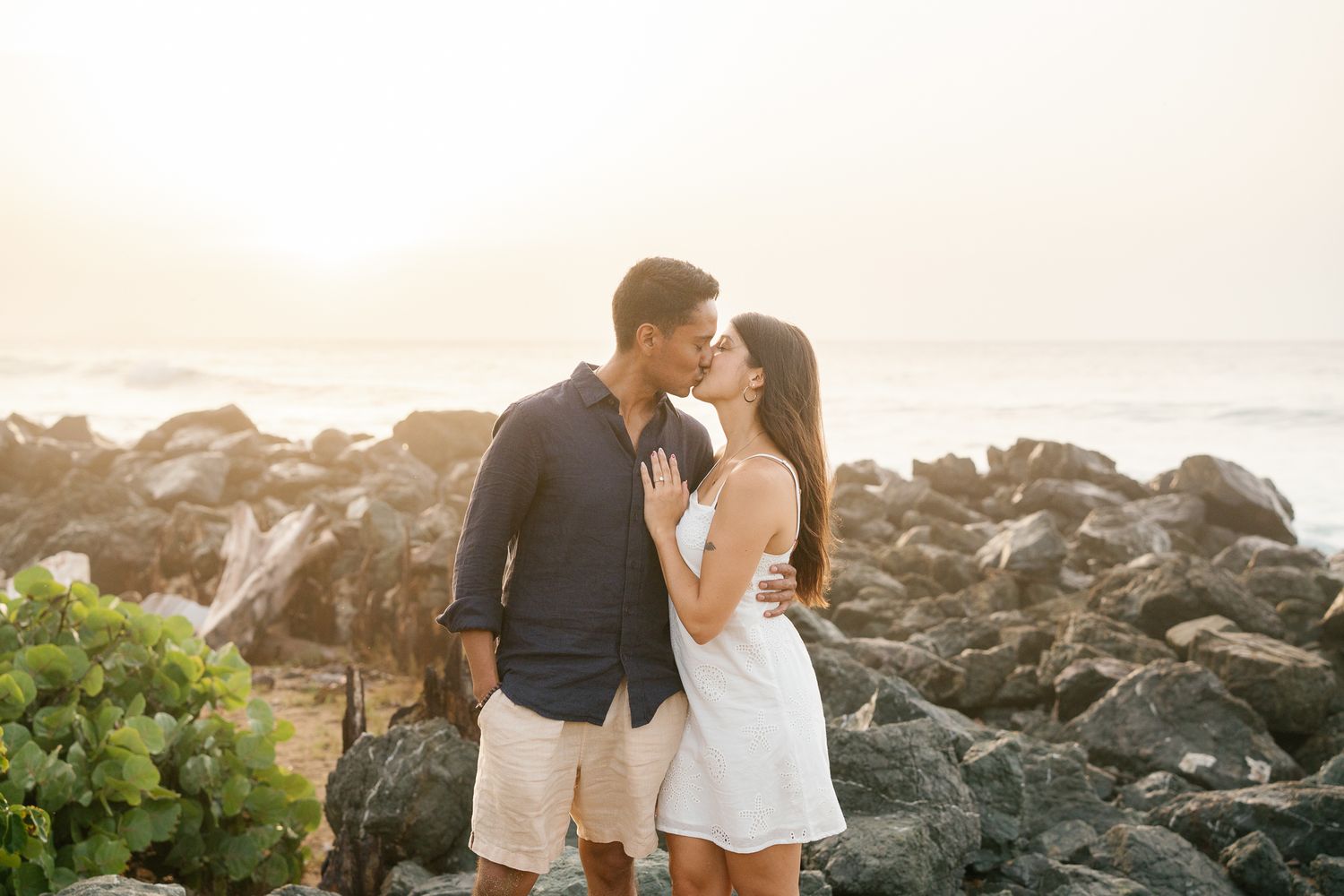 A couple embraces on a rocky beach at sunset with ocean waves crashing in the background.