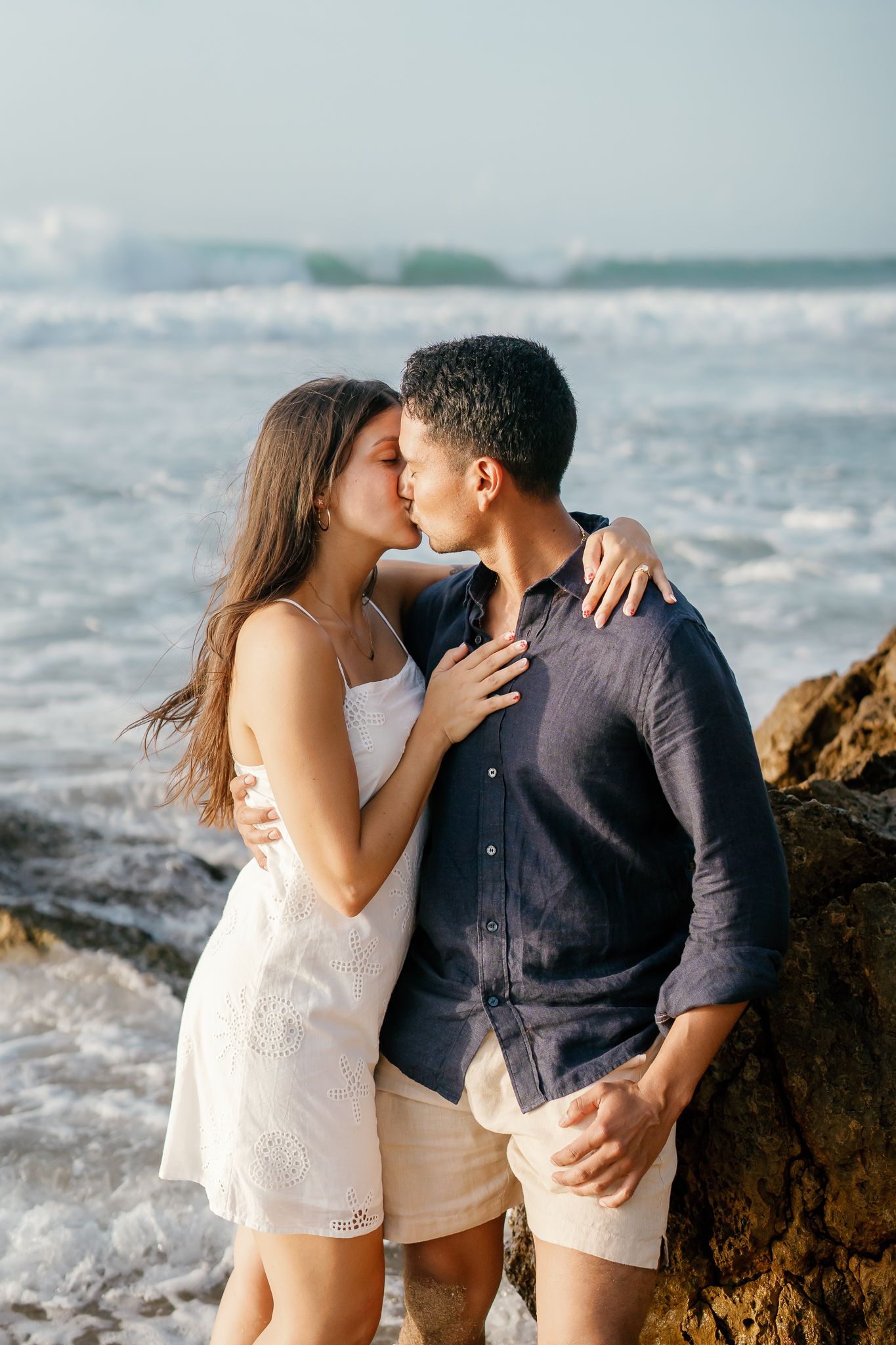 A couple shares romantic kisses on a rocky beach with ocean waves crashing in the background.