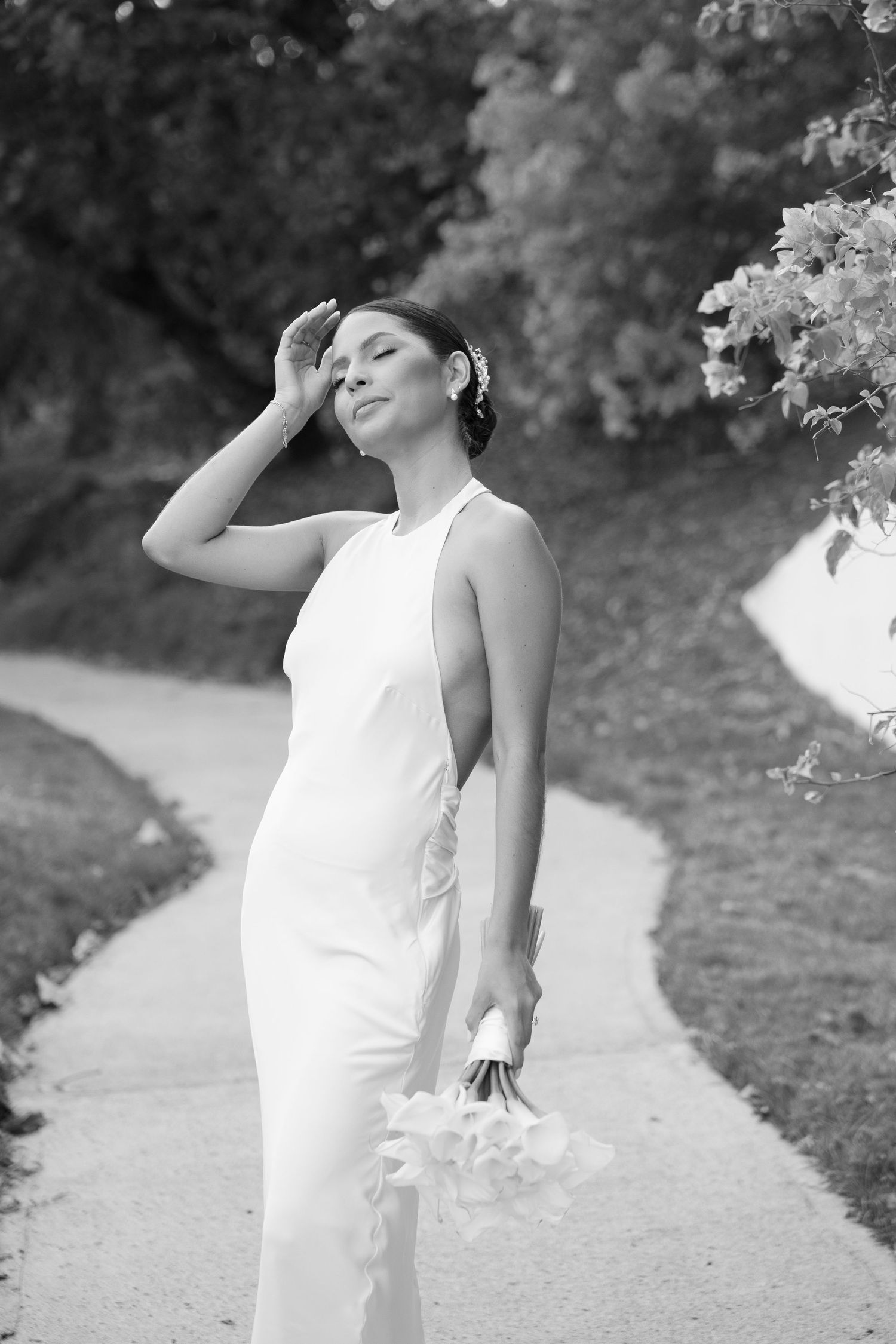 Black and white photo of a bride in a white gown holding calla lilies on a garden path.