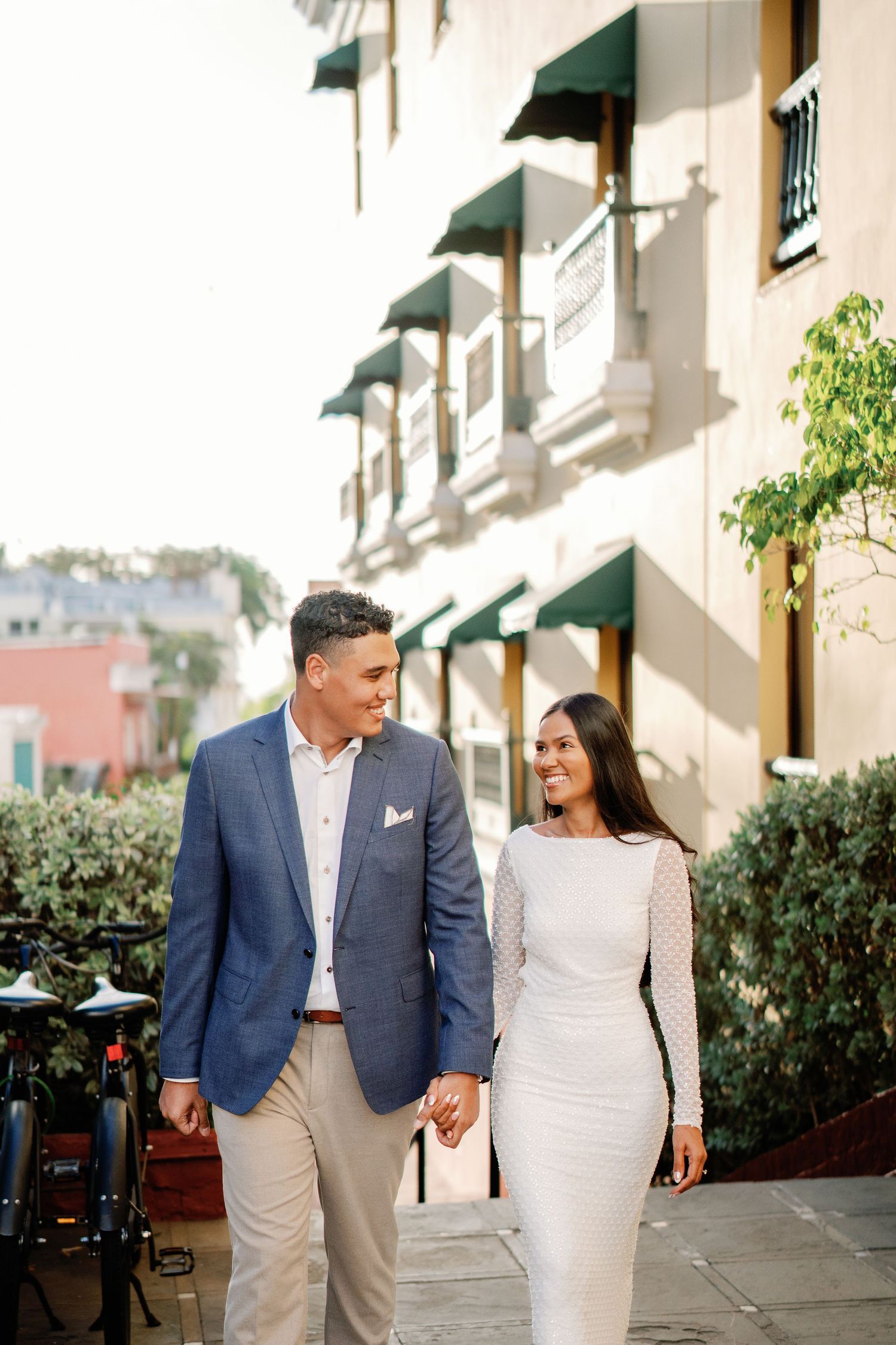 Couple holding hands and smiling while walking along a sunny sidewalk, man in blue blazer, woman in white dress.