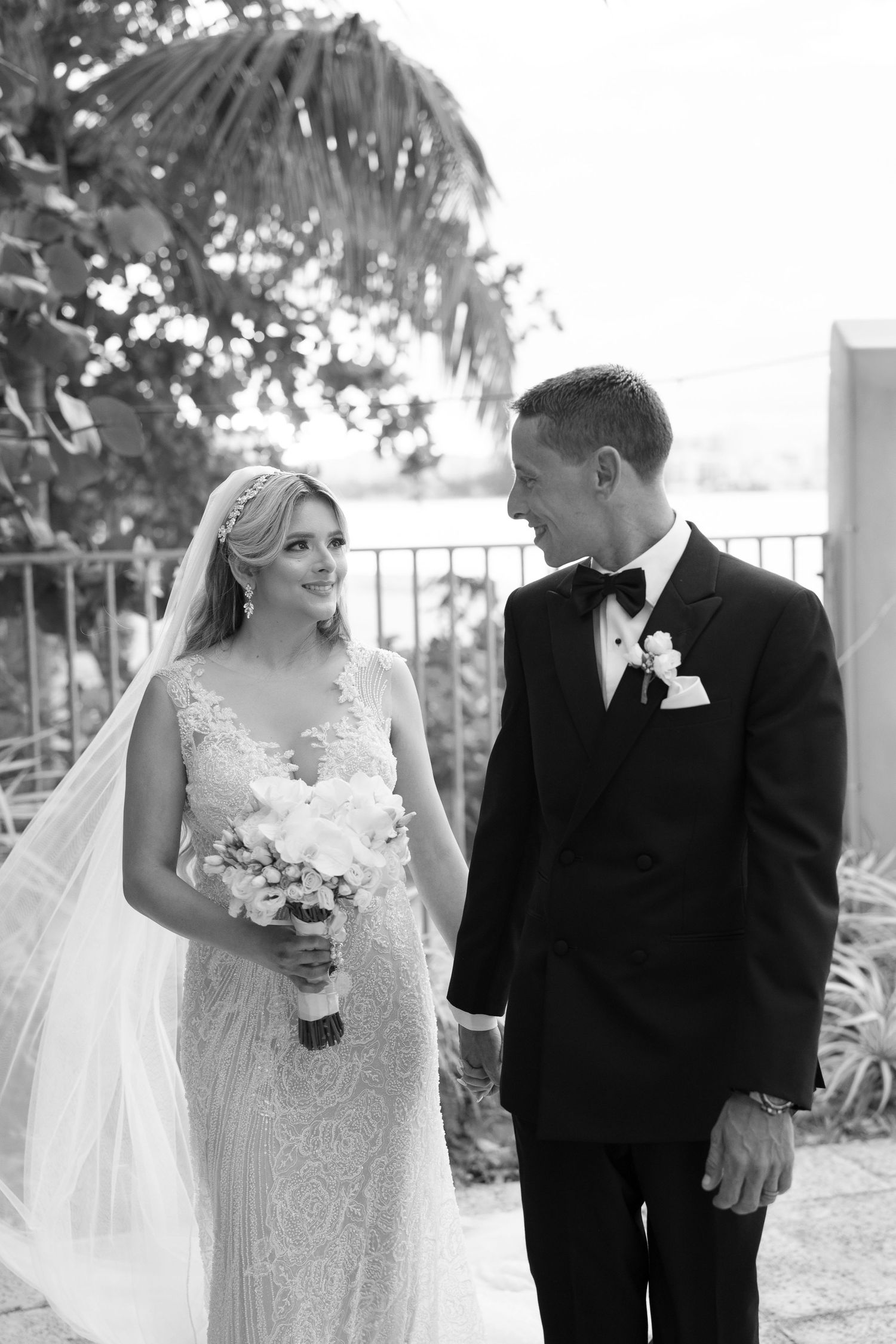 Black and white photo of a smiling bride in lace gown with veil and groom in tuxedo walking together outdoors.