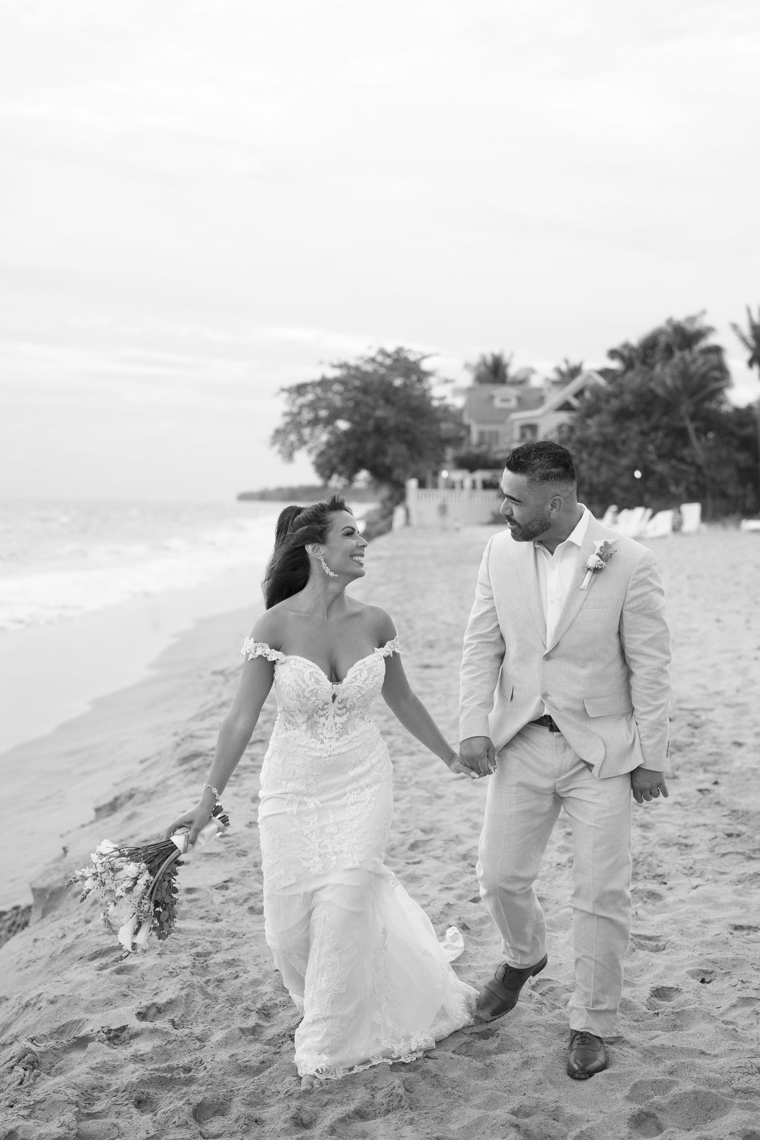 Black and white photo of a bride and groom holding hands, walking on a beach in wedding attire.