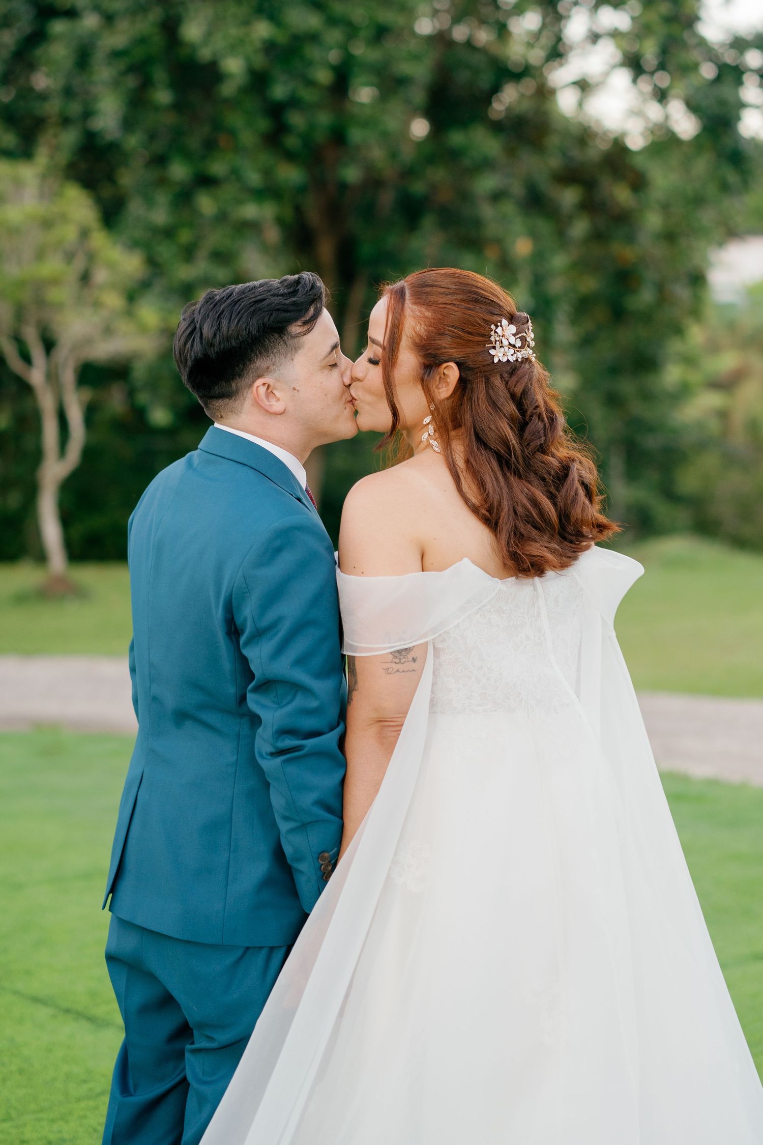 A couple shares a romantic kiss outdoors, groom in blue suit, bride in white off-shoulder gown with auburn hair.