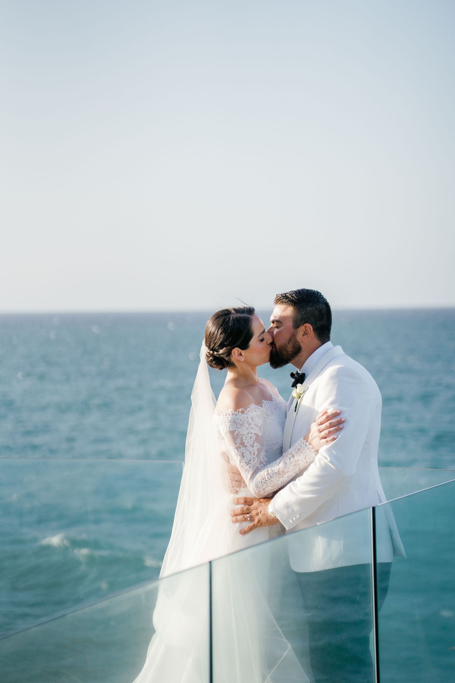 Bride and groom sharing a romantic kiss on a glass balcony overlooking the ocean on their wedding day.