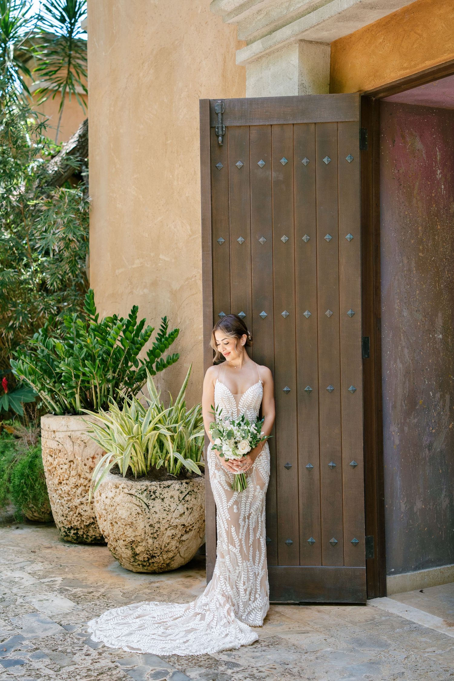 Elegant lace wedding dress with long train shown against rustic wooden door and terra cotta wall with potted plants.