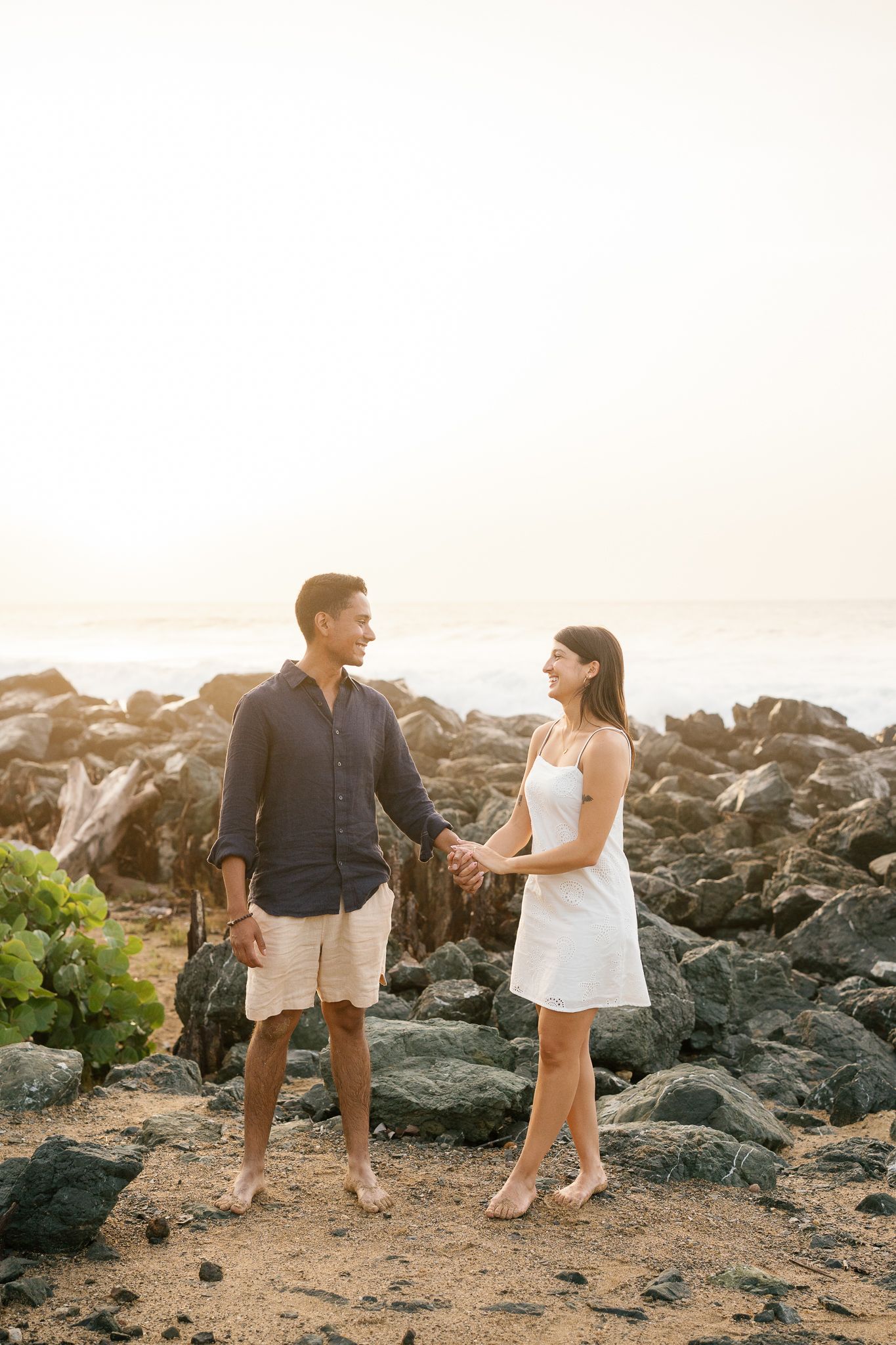 A couple shares a romantic moment at sunset while standing on rocky coastal terrain.