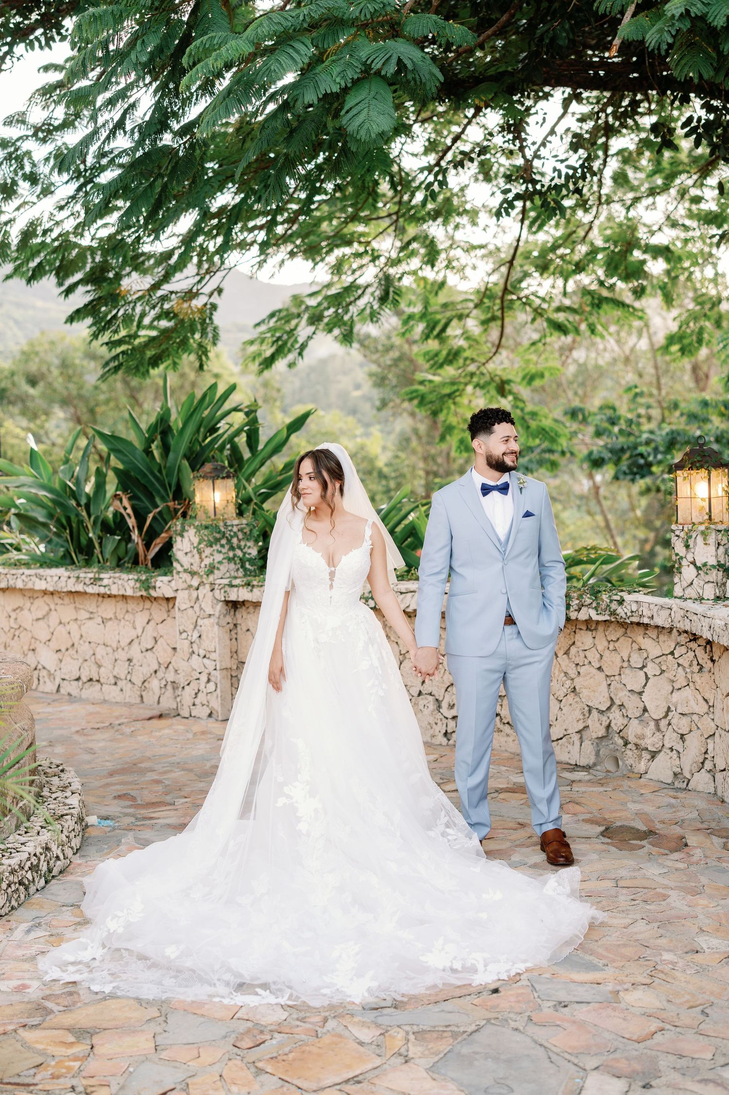 Bride in lace gown with veil and groom in light blue suit hold hands outdoors at a lush garden wedding venue.