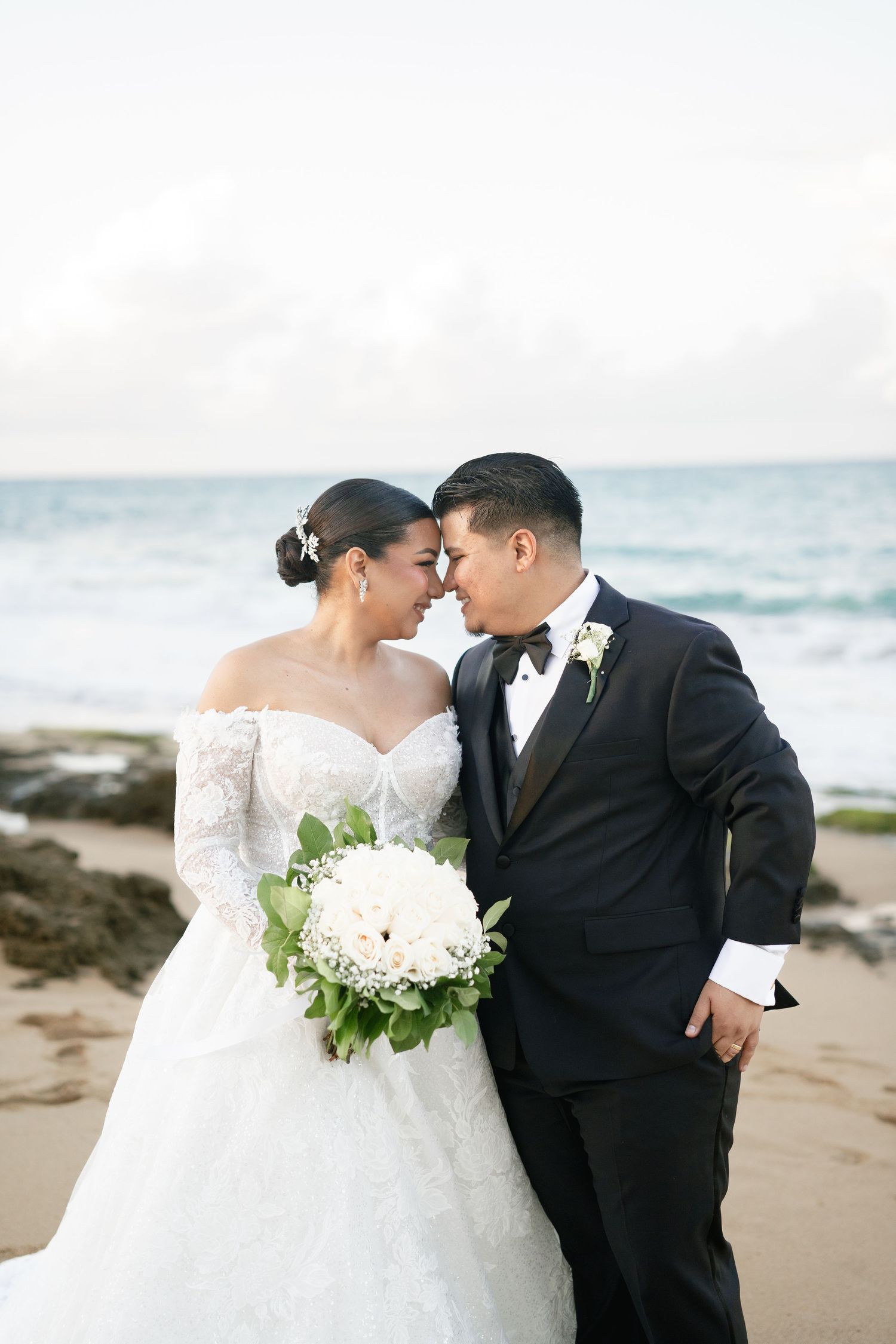 Bride and groom share a romantic forehead touch on a sandy beach, bride holding white floral bouquet in lace gown.