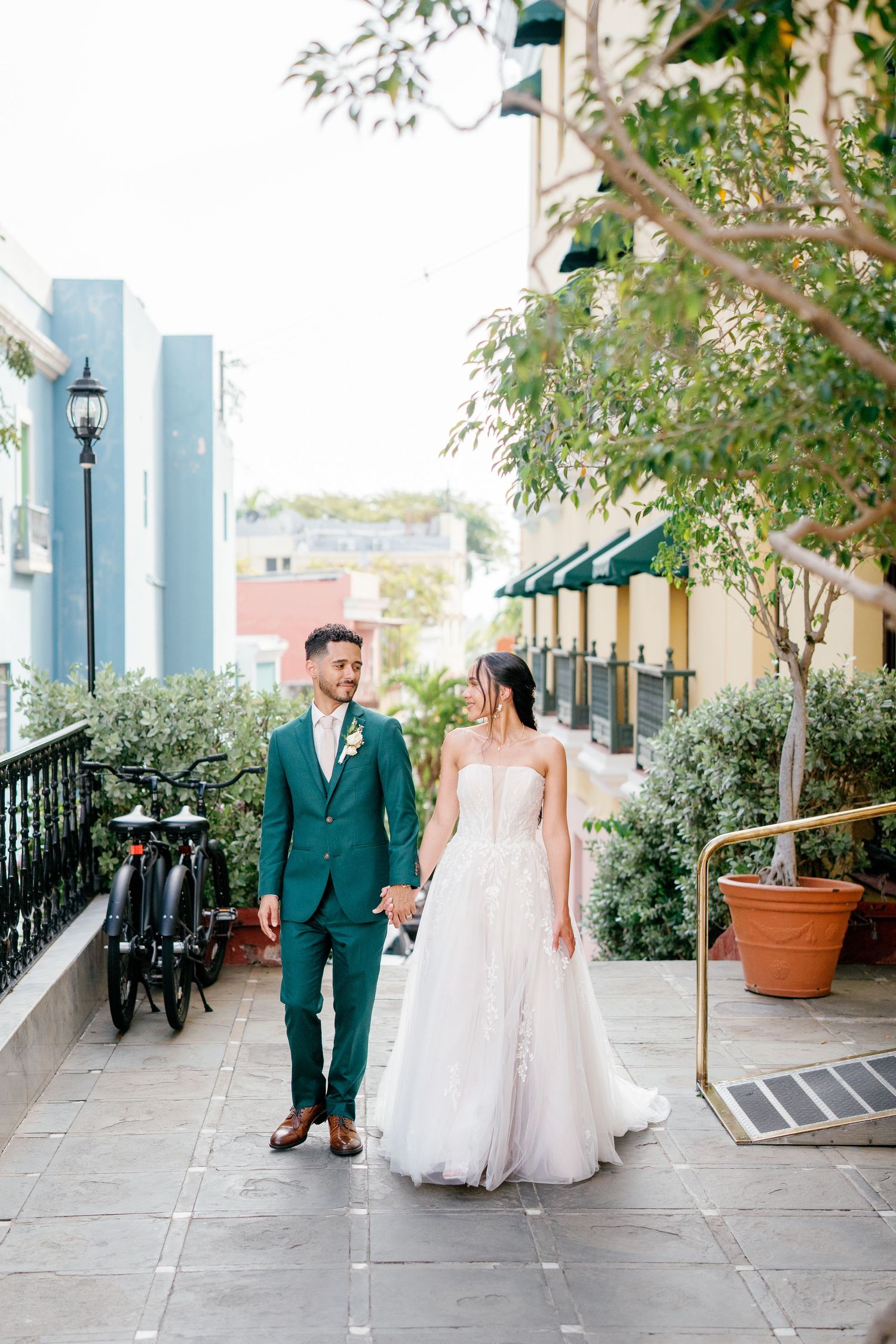 Bride in white gown and groom in green suit walk hand in hand along a charming outdoor corridor.