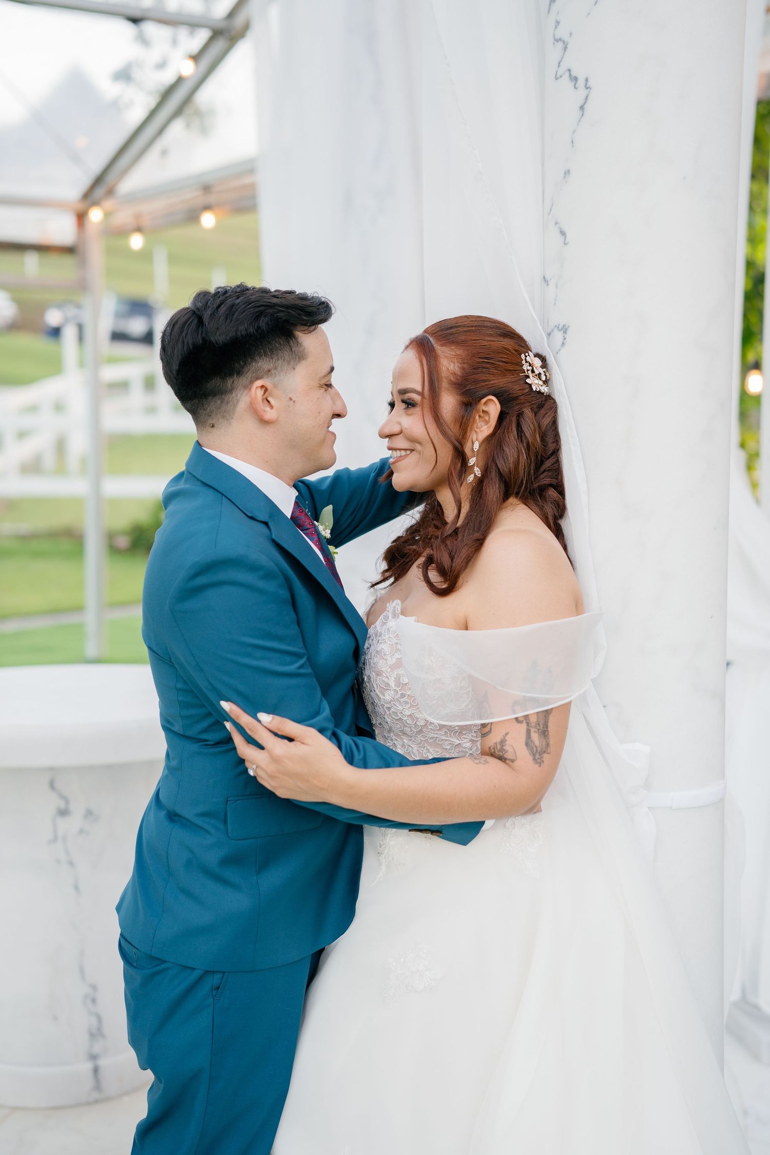 Bride and groom share a romantic moment by a marble column, dressed in a blue suit and white wedding gown.