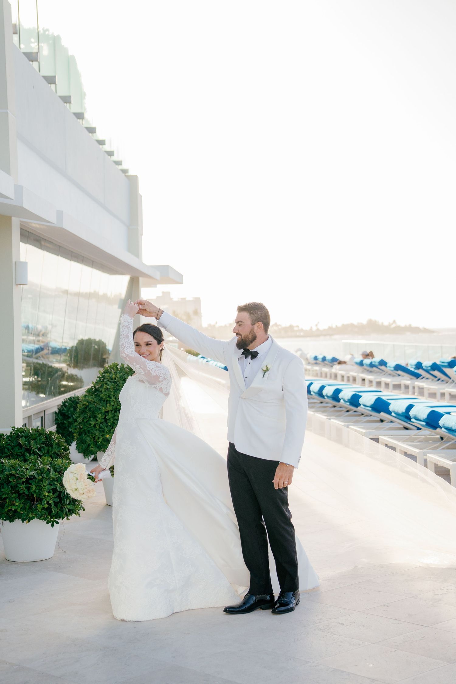 Groom twirls bride in a lace wedding gown on a bright waterfront terrace with blue lounge chairs in the background.