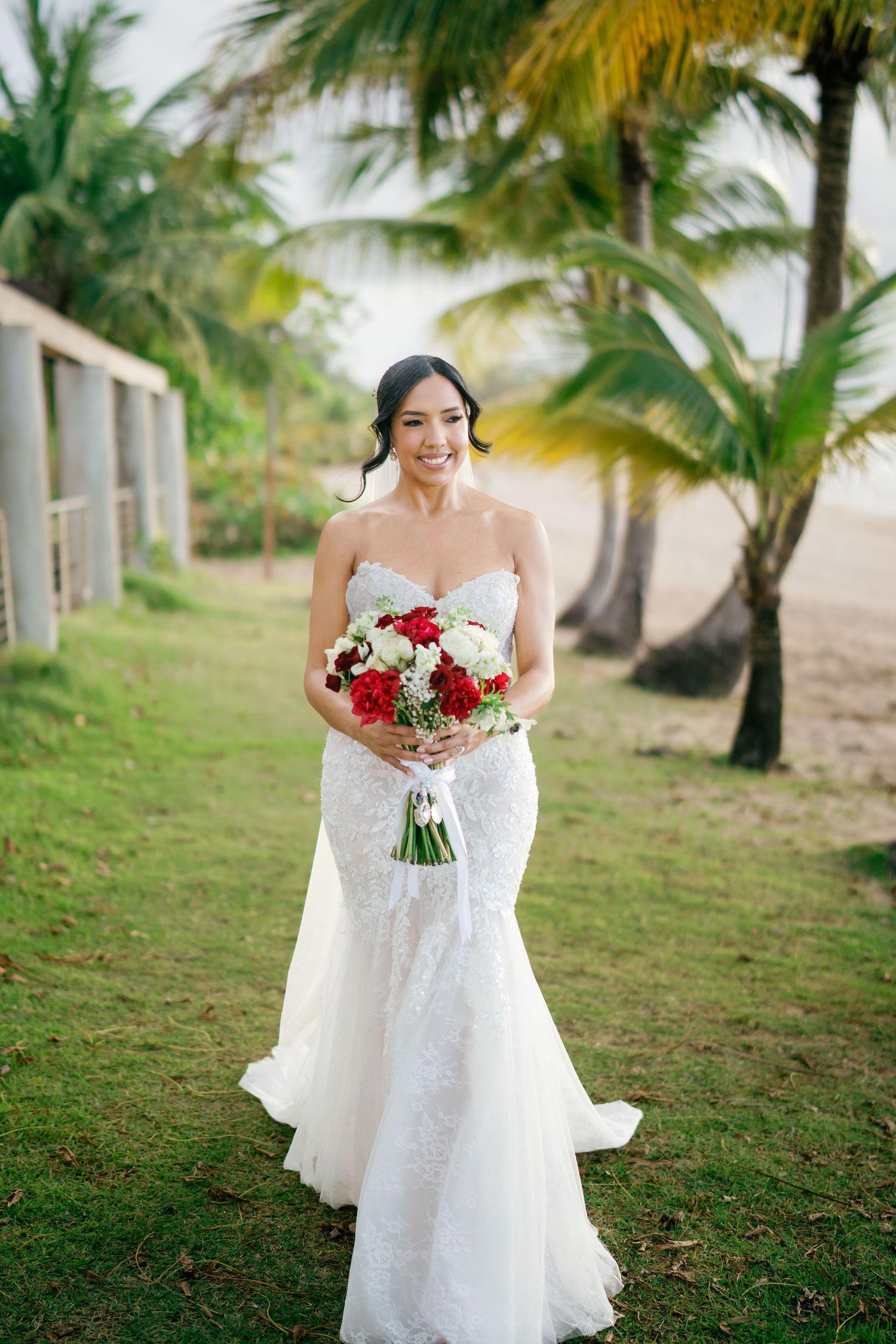 Bride in white lace mermaid gown holding red and white bouquet standing among tropical palm trees.