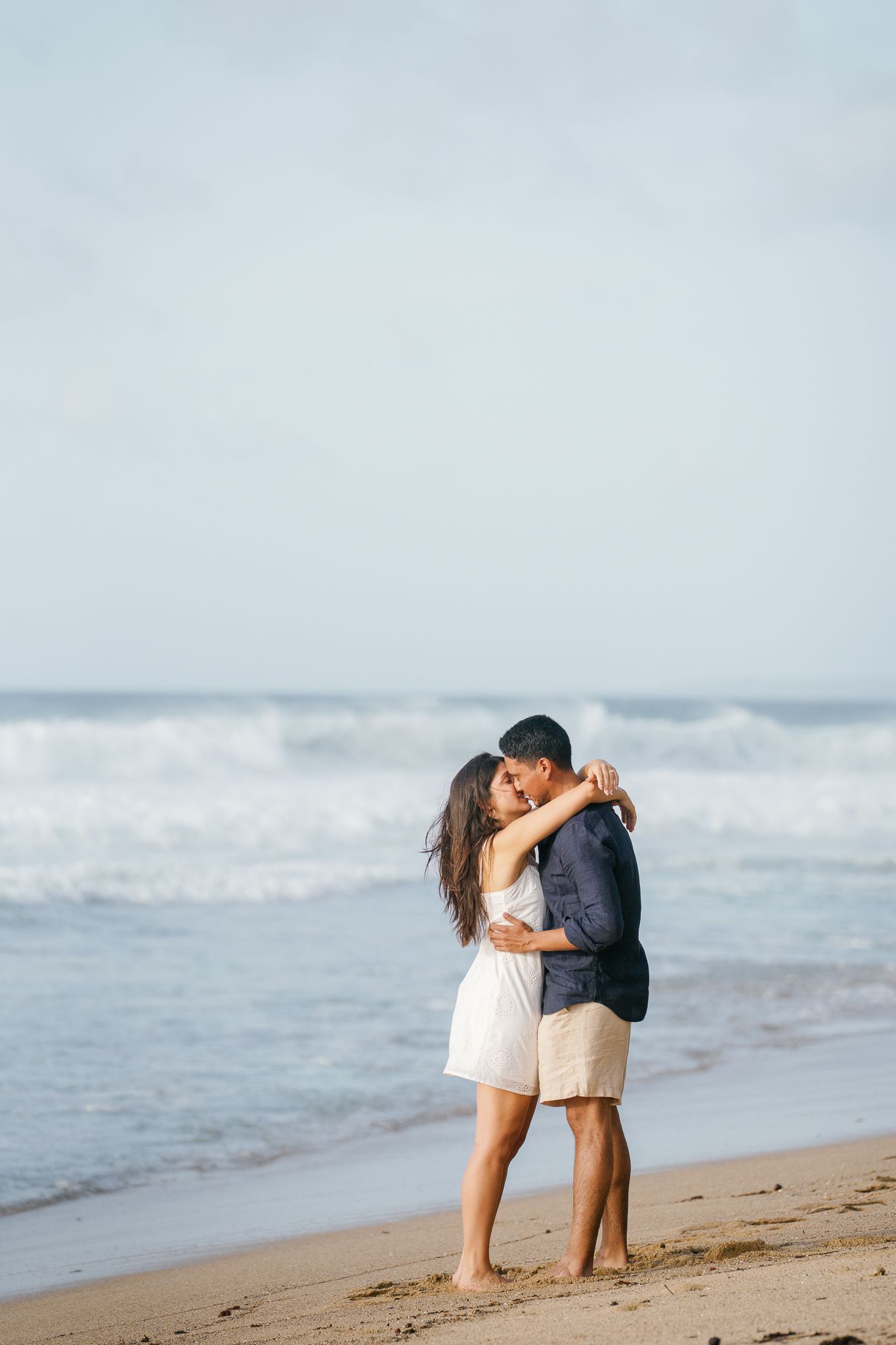 A couple embraces romantically on a sandy beach with ocean waves in the background.