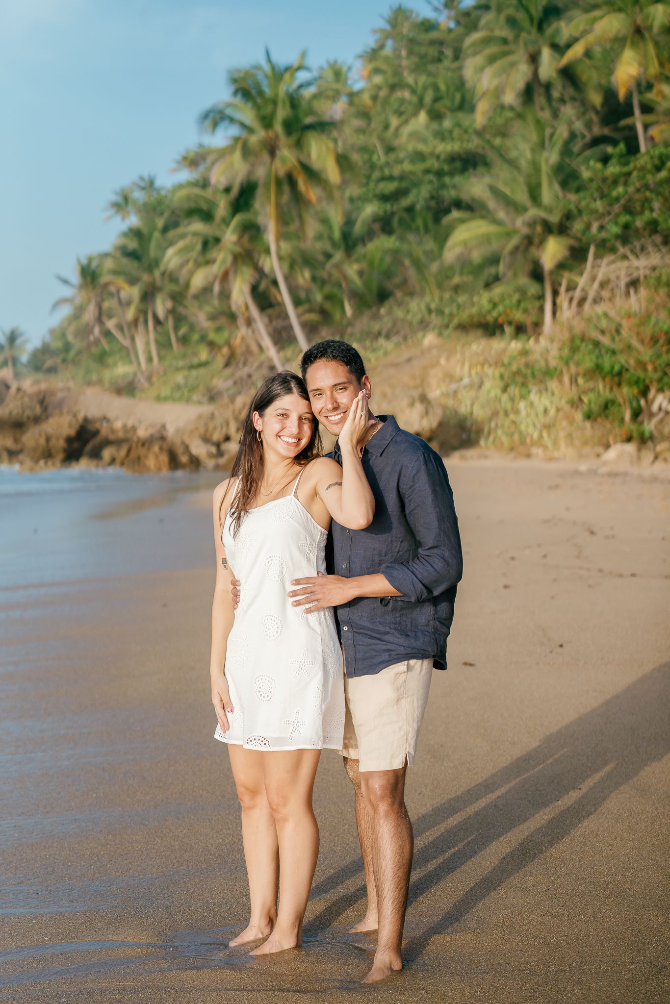 A couple embraces on a tropical beach with palm trees and coastline in the background during golden hour.
