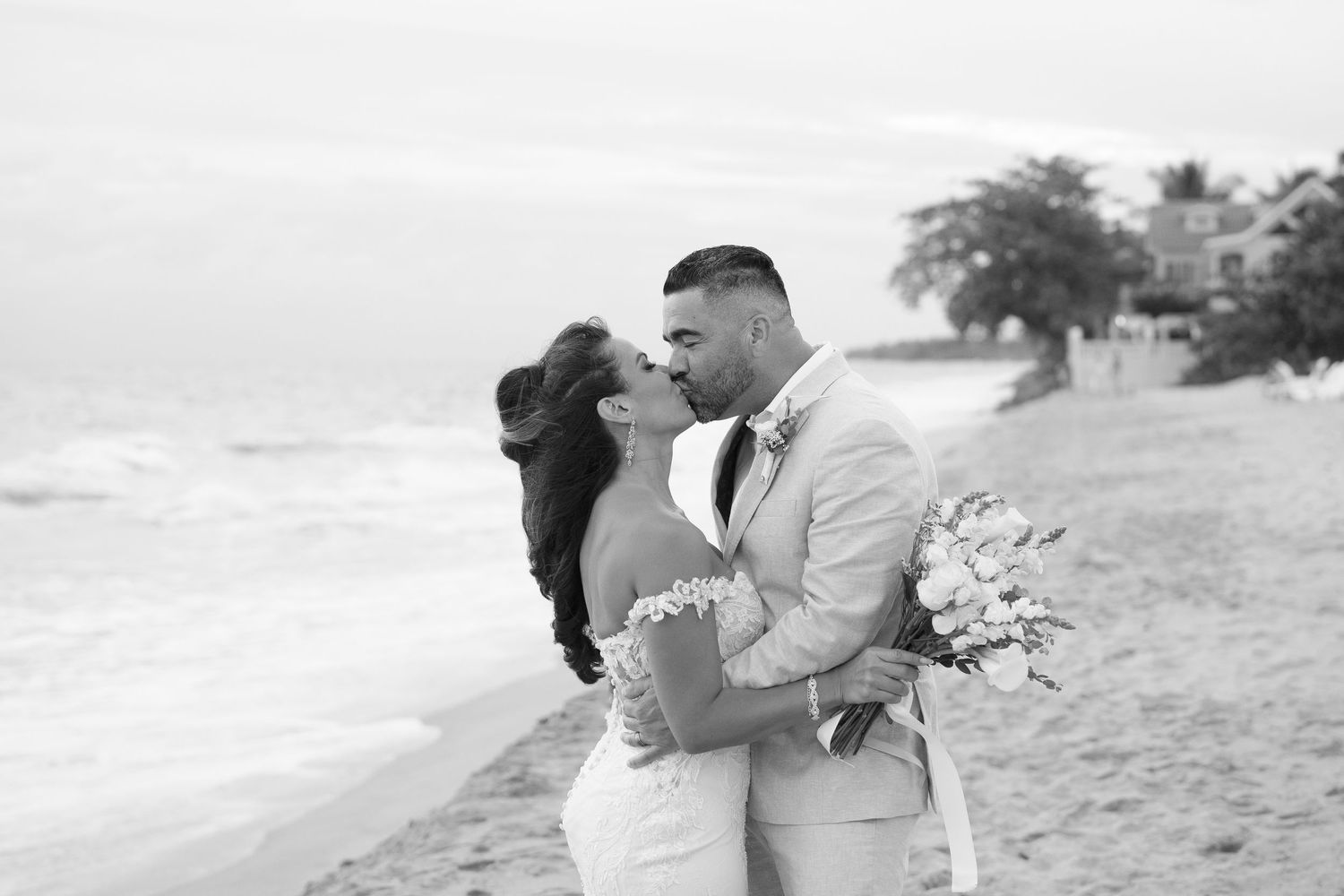 Black and white photo of a couple sharing a romantic kiss on a beach in wedding attire, holding a bouquet.