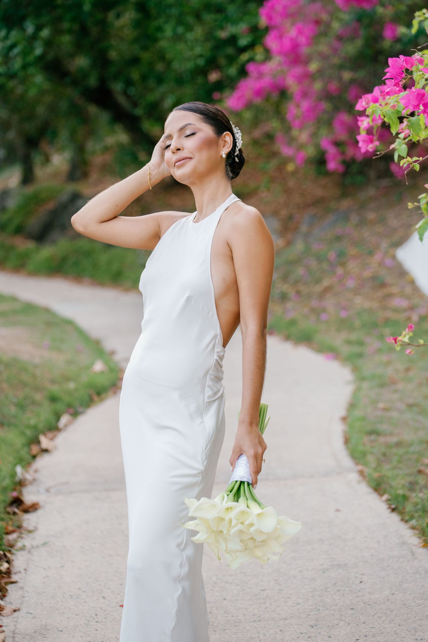Bride in sleek white halter gown holding calla lily bouquet on garden path with pink bougainvillea blooms.