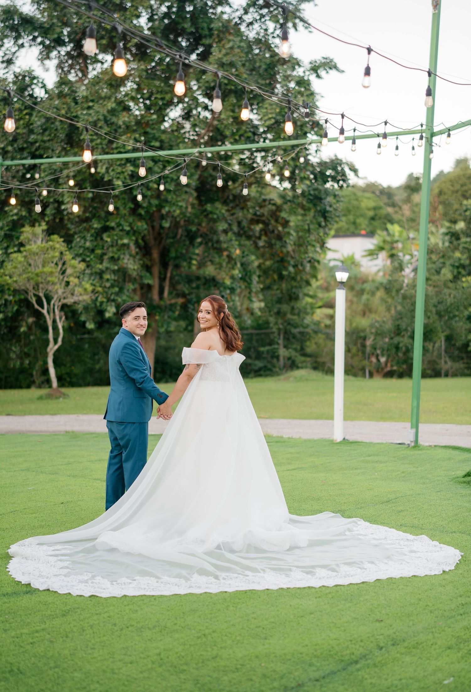 Bride in off-shoulder gown and groom in blue suit hold hands on green lawn under string lights at outdoor wedding.