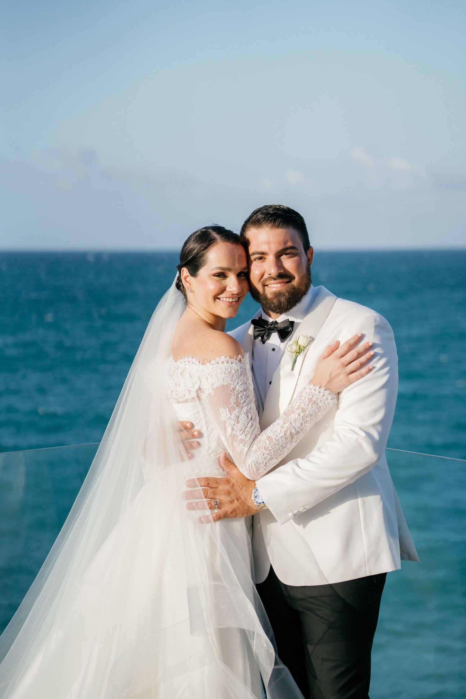 Bride and groom embrace on oceanfront balcony, she in lace off-shoulder gown with veil, he in white tuxedo jacket.