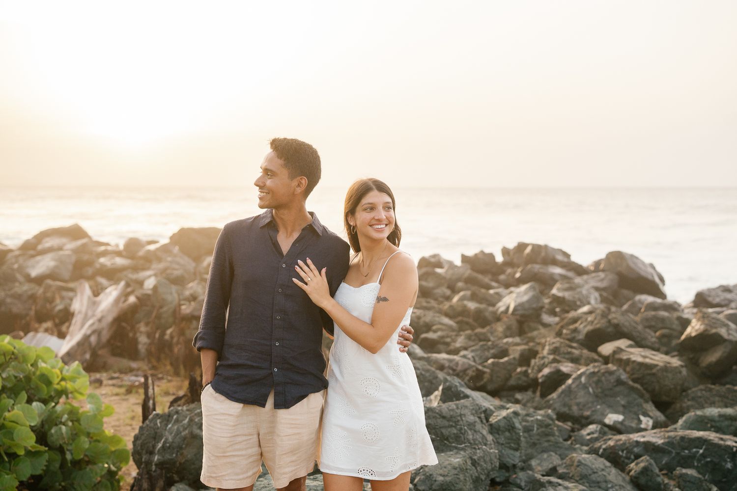 A couple shares a romantic moment at sunset on a rocky beach shoreline, embracing and laughing together.